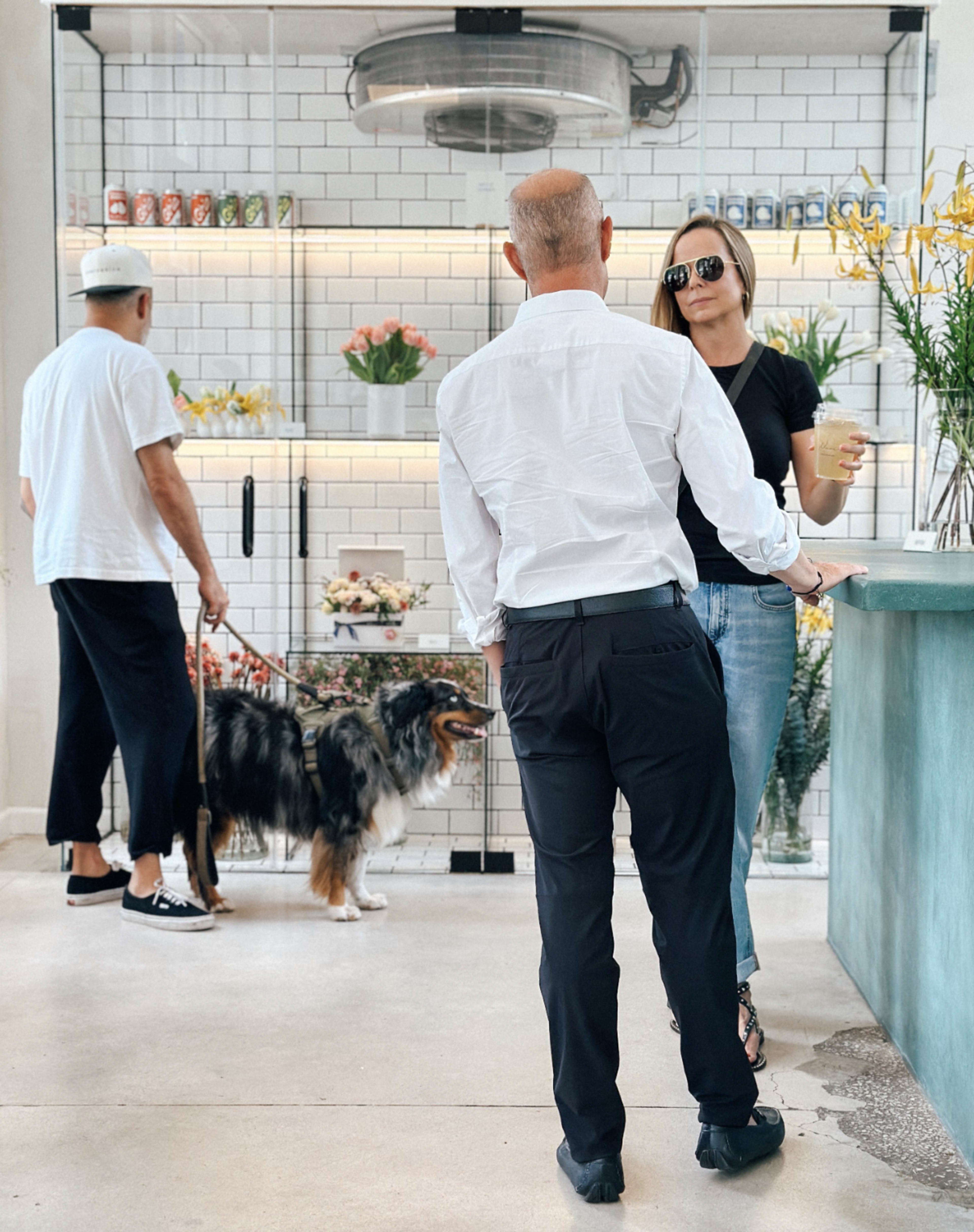 Two people stand at a counter while a man walks a dog in a shop decorated with flowers and drinks on display.
