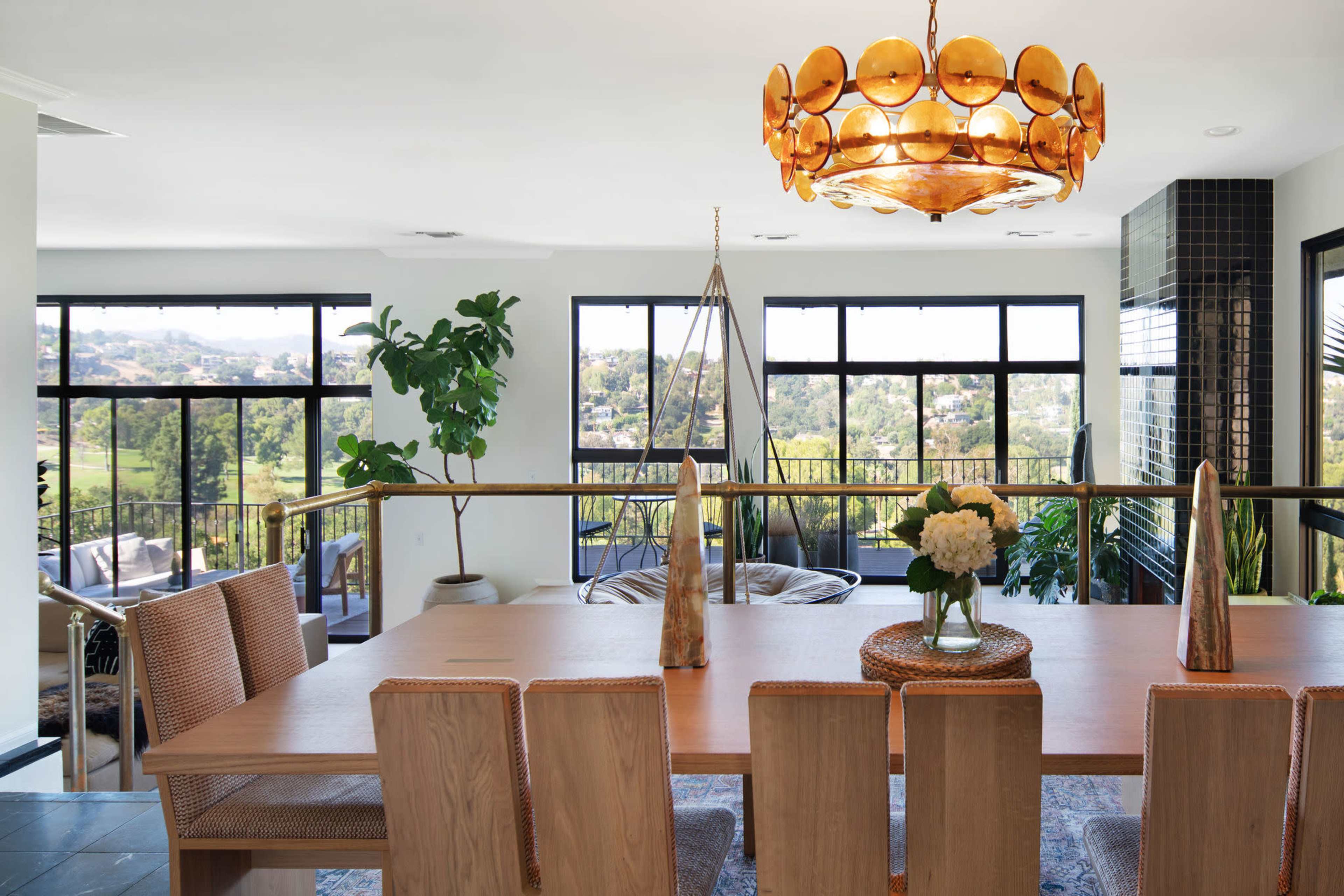 A modern dining area with a long wooden table, woven chairs, and a decorative chandelier, featuring large windows that overlook a scenic view.