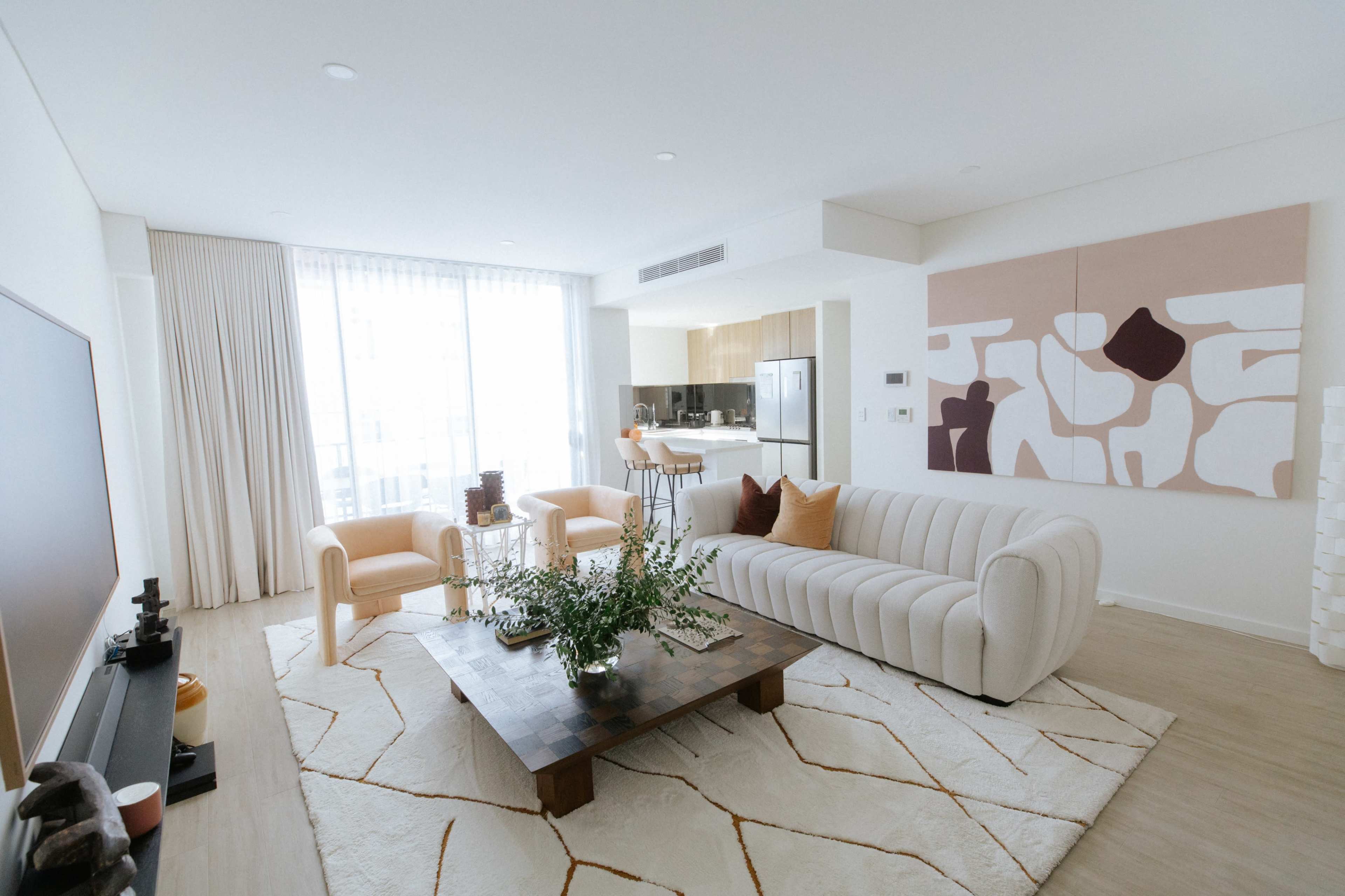 A modern living room features a beige sofa, two orange chairs, a wooden coffee table with a floral arrangement, and large windows with light-colored curtains.