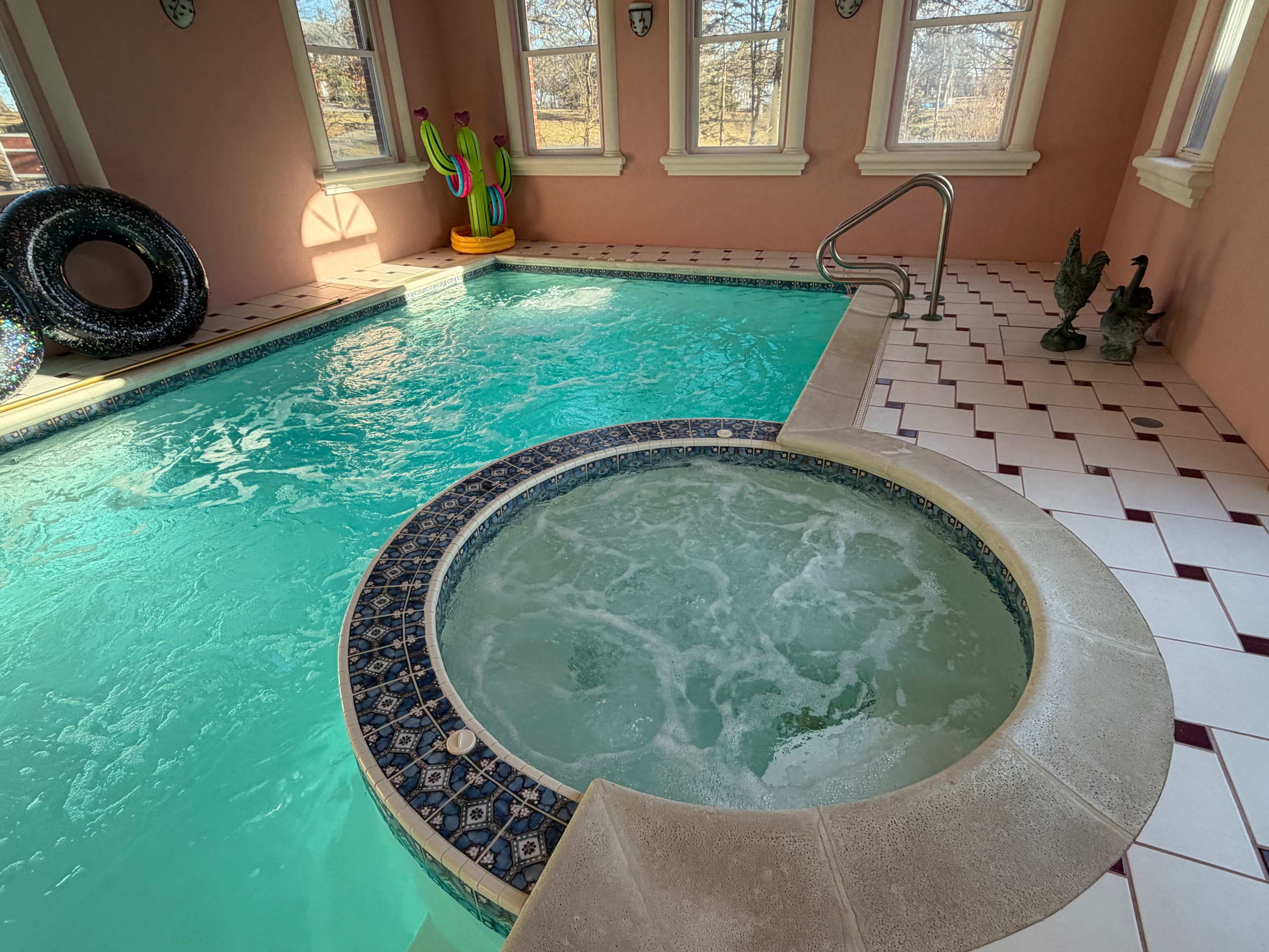 The image shows an indoor pool area featuring a large swimming pool with a circular hot tub integrated into the design, surrounded by pink walls and decorative windows.
