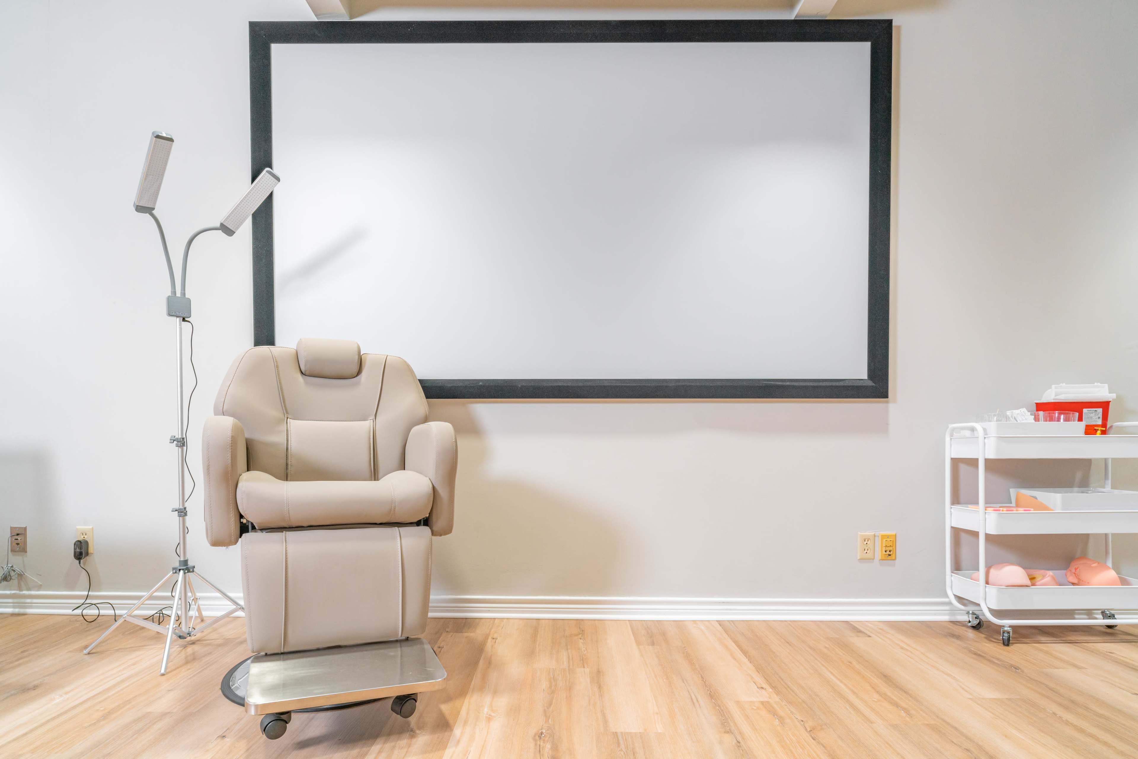 The image shows a beige reclining chair positioned next to a blank wall framed by a dark border, with a floor lamp and a mobile cart containing supplies nearby.