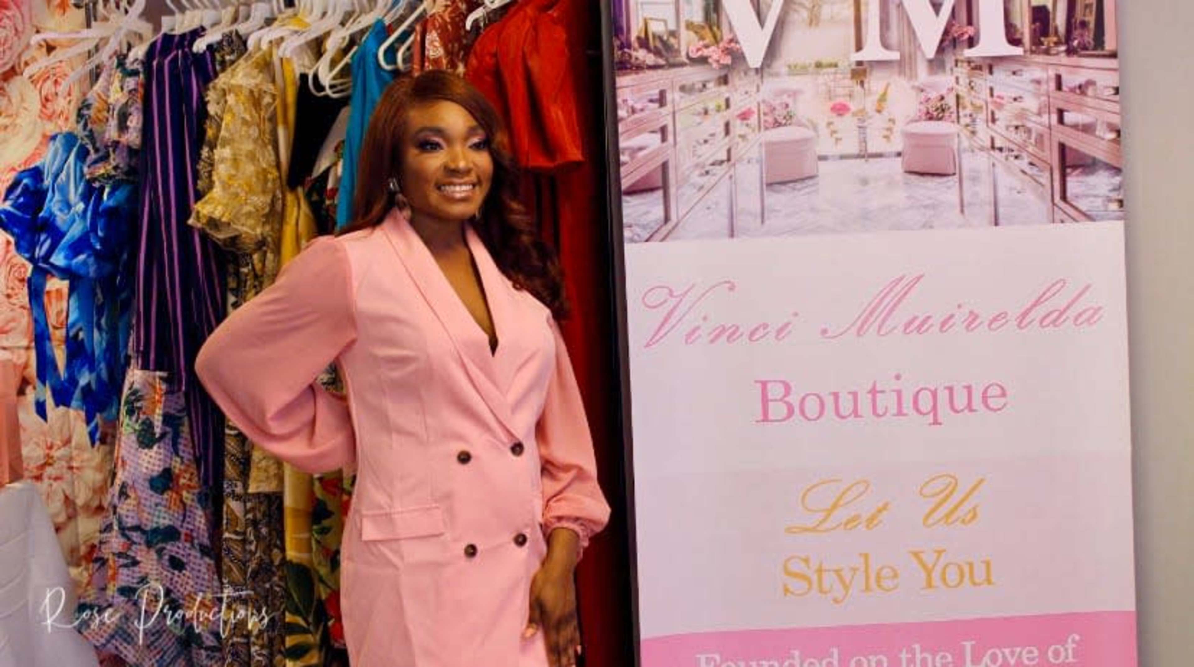 A woman wearing a pink outfit poses next to a sign for a boutique named "Vinci Muirelda."