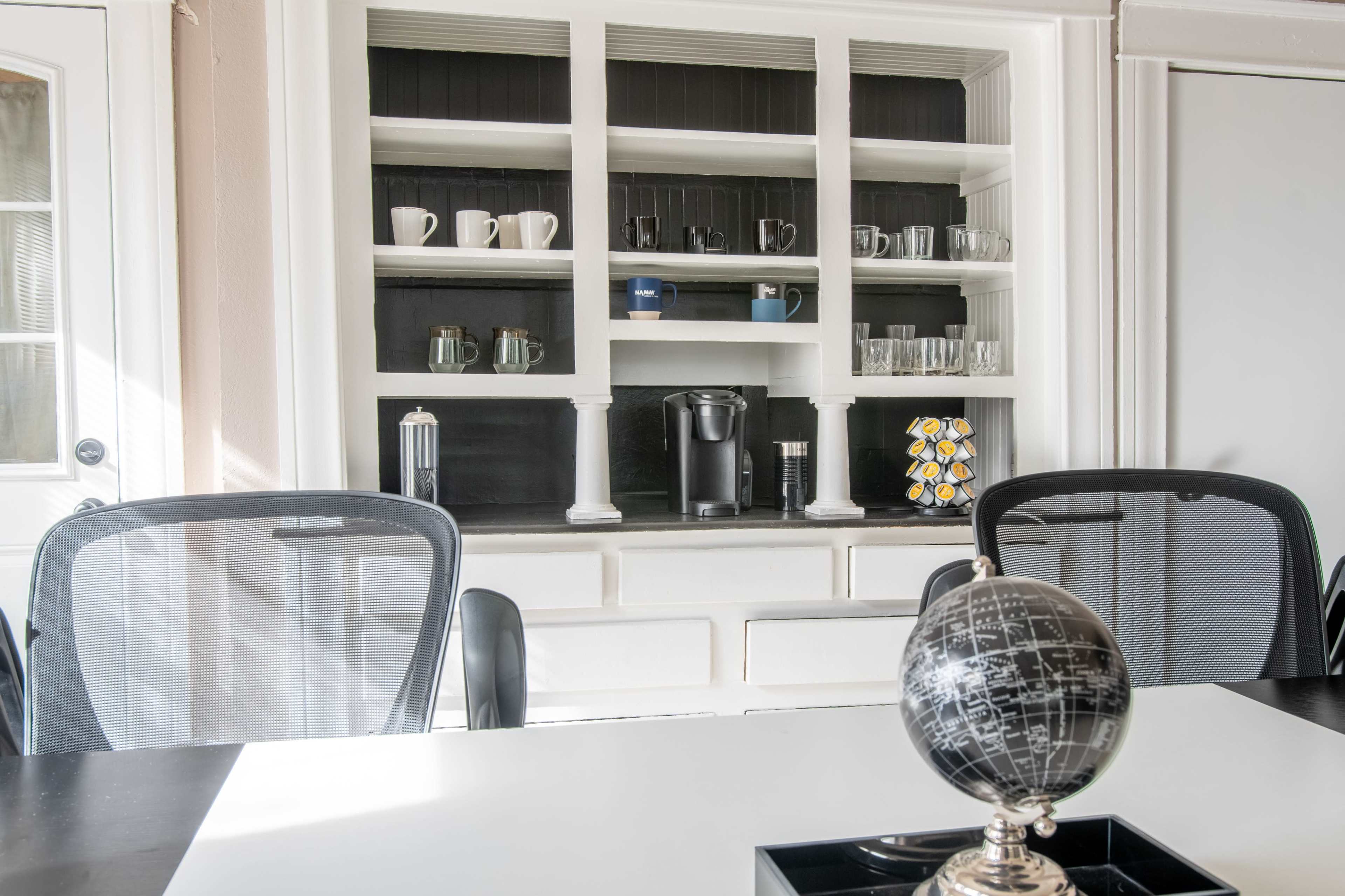 A modern kitchen area with a white shelving unit displaying glassware and a coffee maker, set against a backdrop of a black wall.