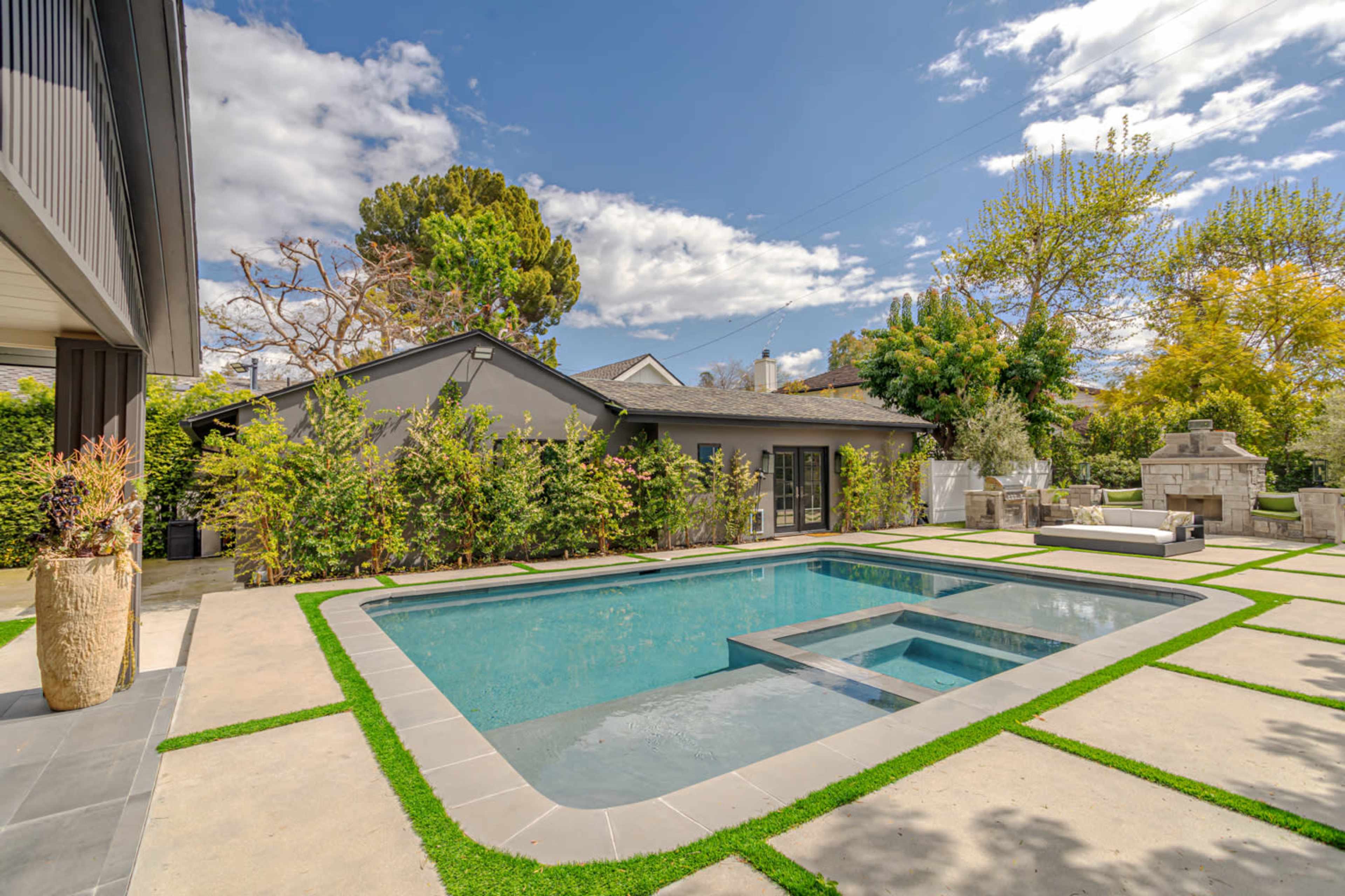 The image shows a modern backyard featuring a rectangular swimming pool with a spa, surrounded by landscaped greenery and concrete pathways.