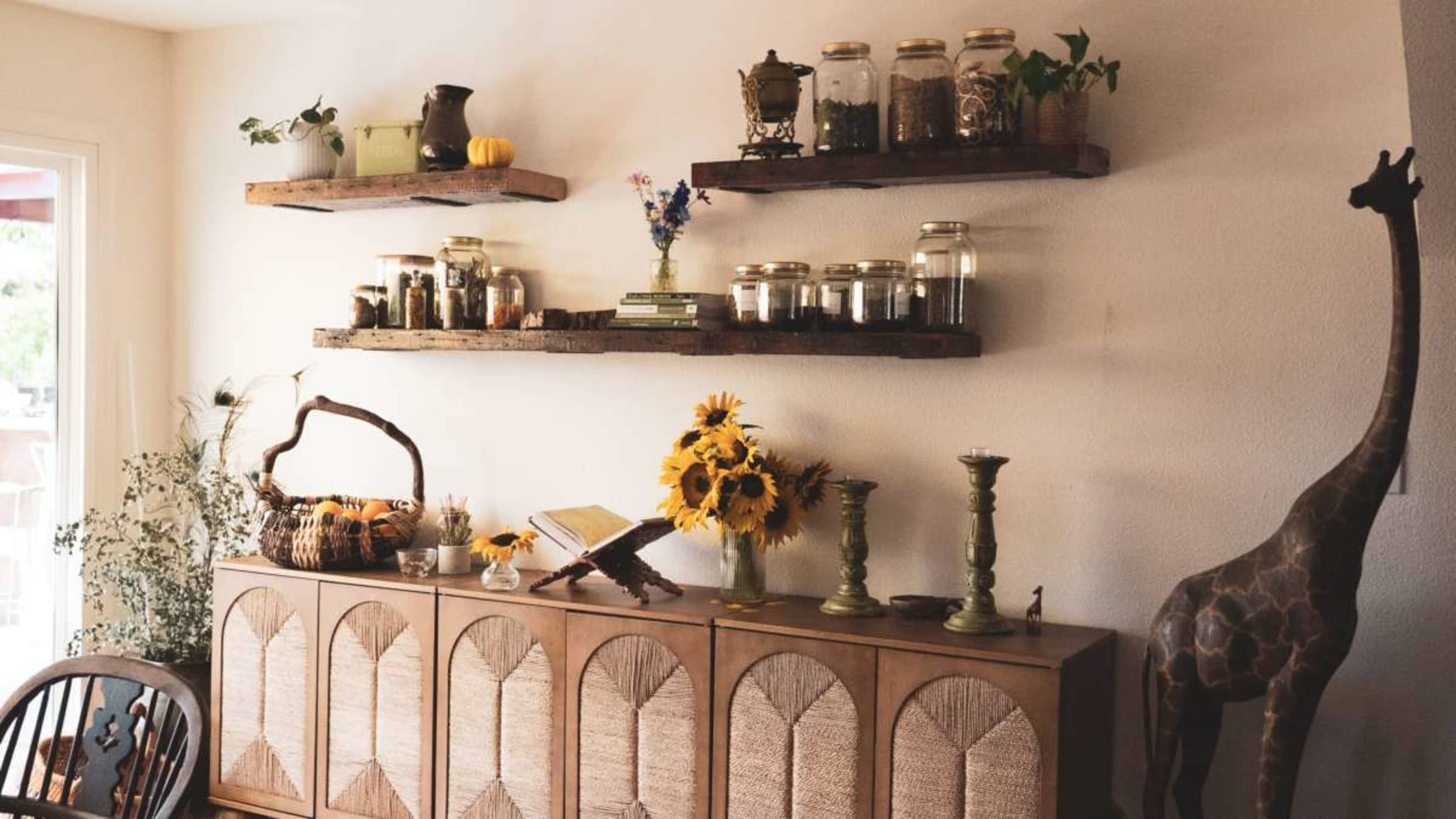 The image shows a cozy interior with wooden shelves displaying jars of herbs, flowers, and decorative items, alongside a cabinet adorned with sunflowers and various artifacts.