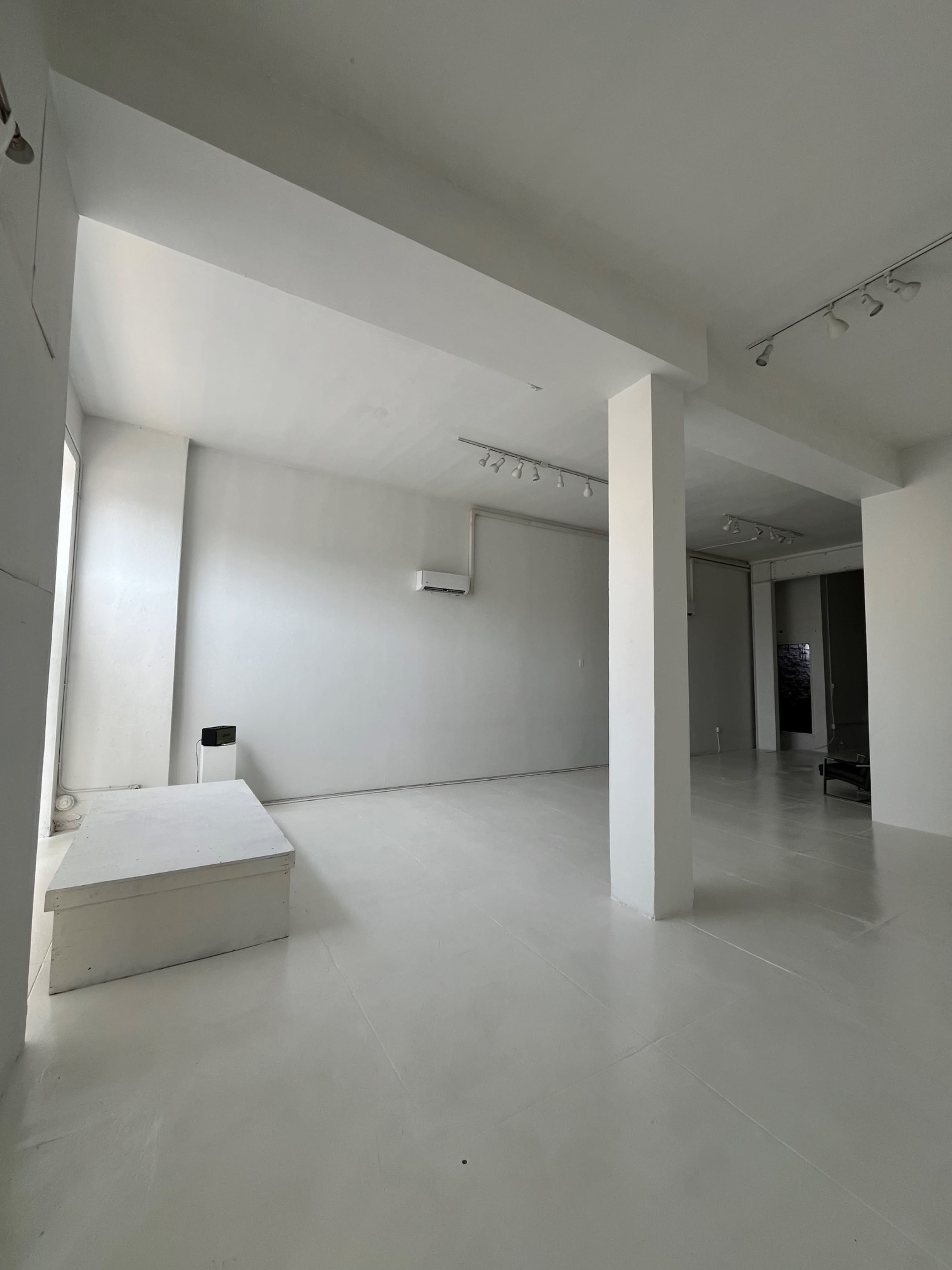 The image shows an empty, minimalist room with white walls and a light-colored floor, featuring a small raised platform and an air conditioning unit on one side.