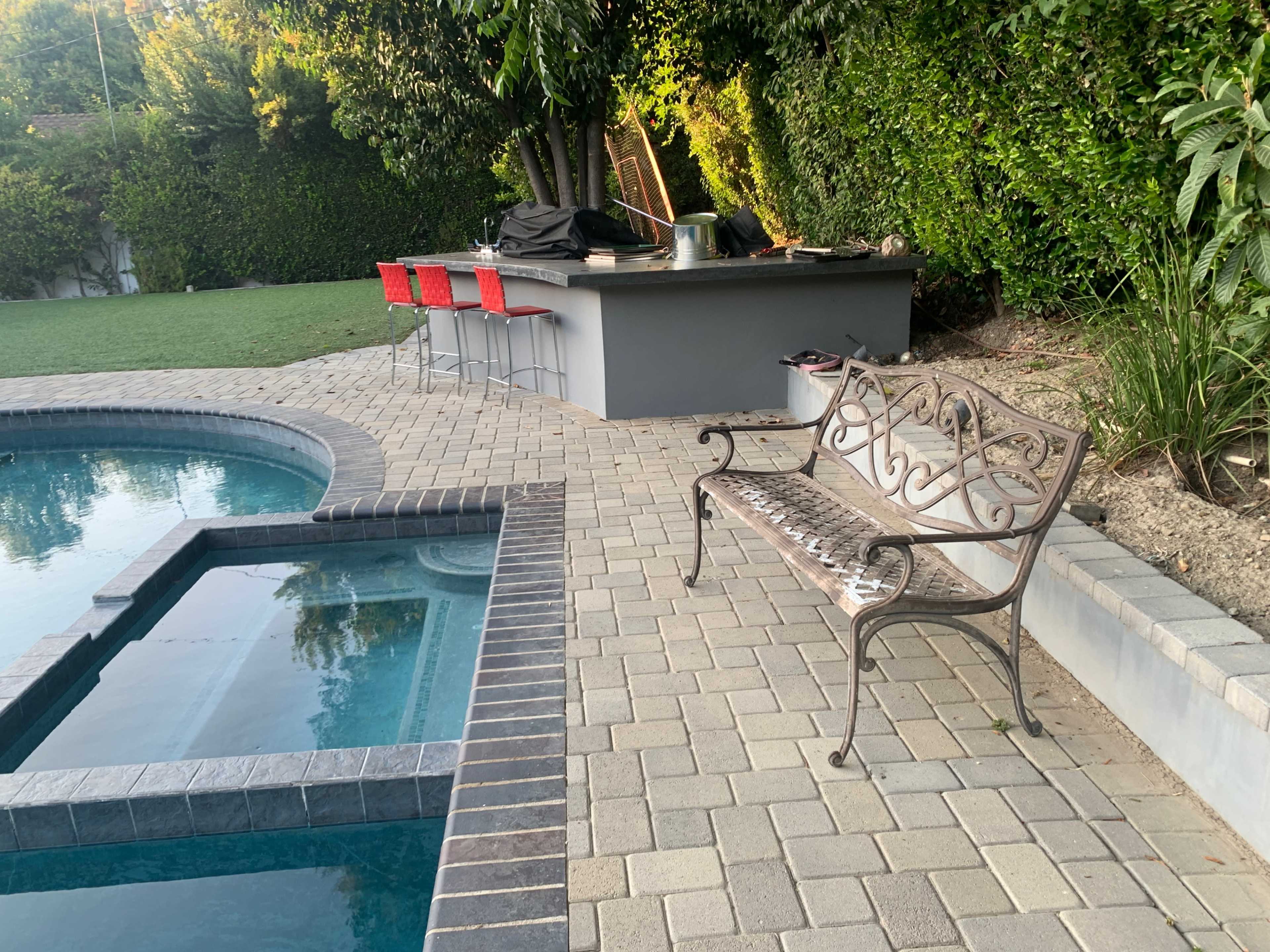 The image shows a stone patio with a pool on one side, a bench nearby, and a barbecue area with red stools in the background.