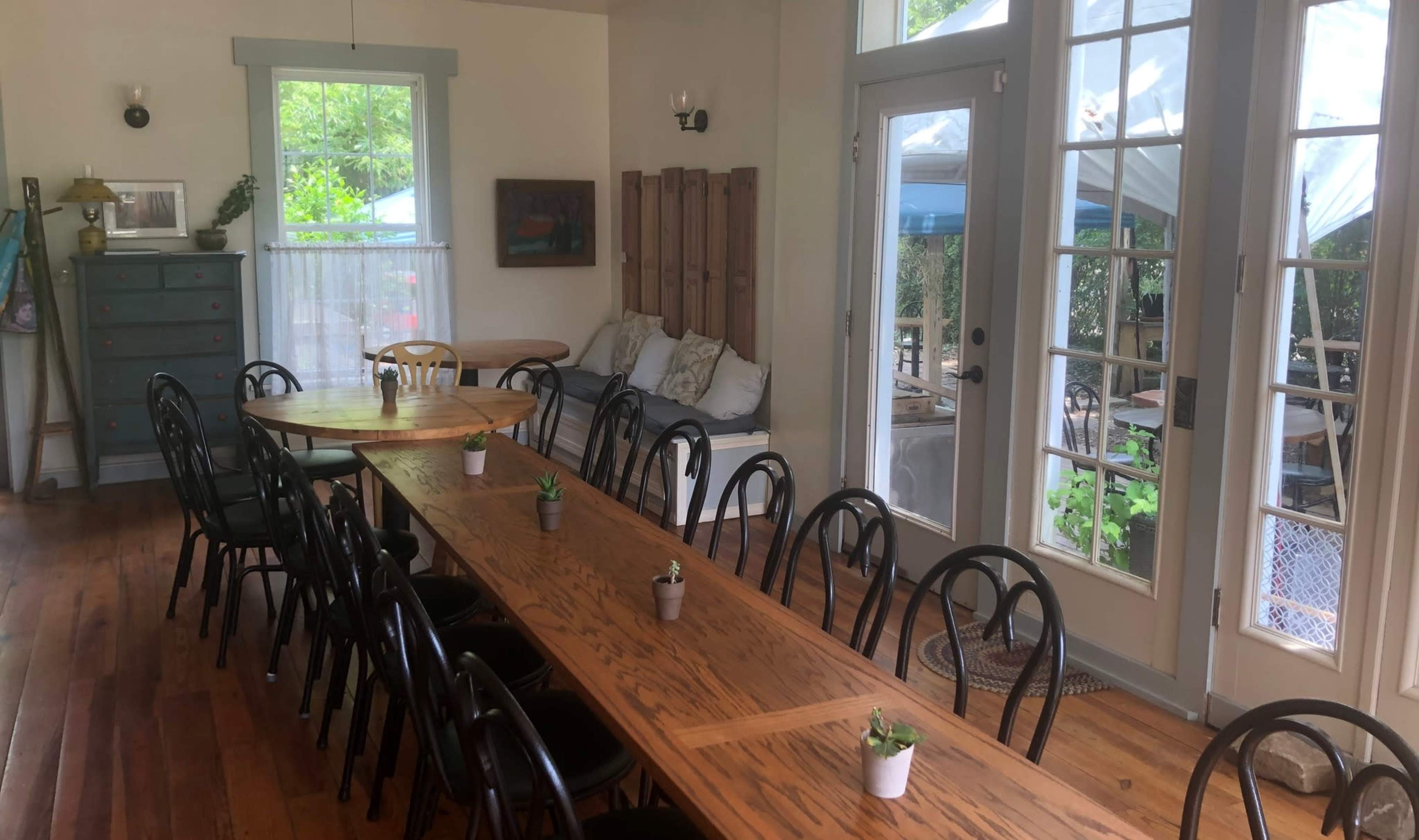 A dining area with a long wooden table surrounded by black chairs, large windows letting in natural light, and a door leading to an outdoor space with seating.