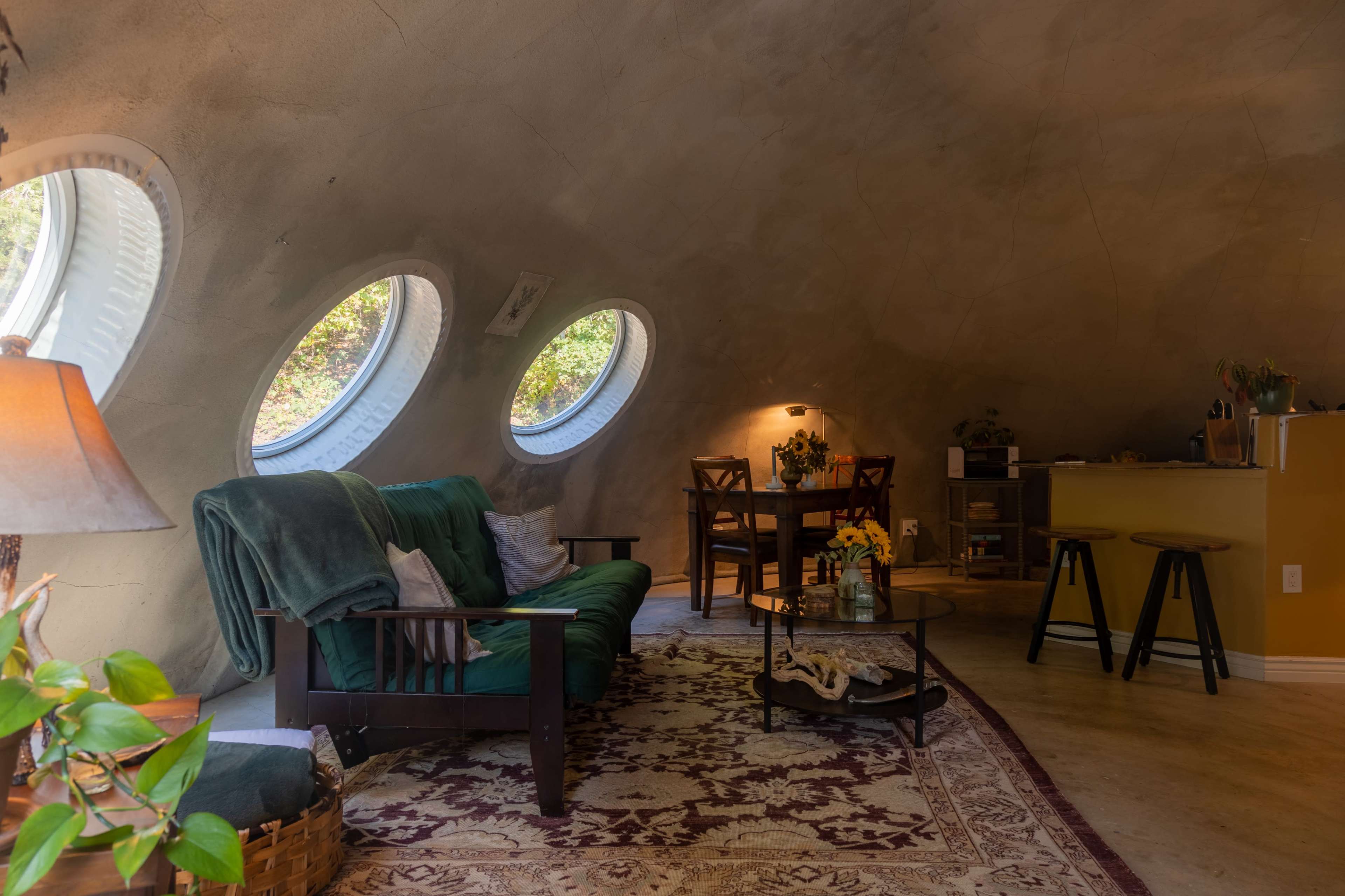 The image shows a bright, dome-shaped living space featuring a green couch, a wooden dining table, and large circular windows that offer a view of greenery outside.