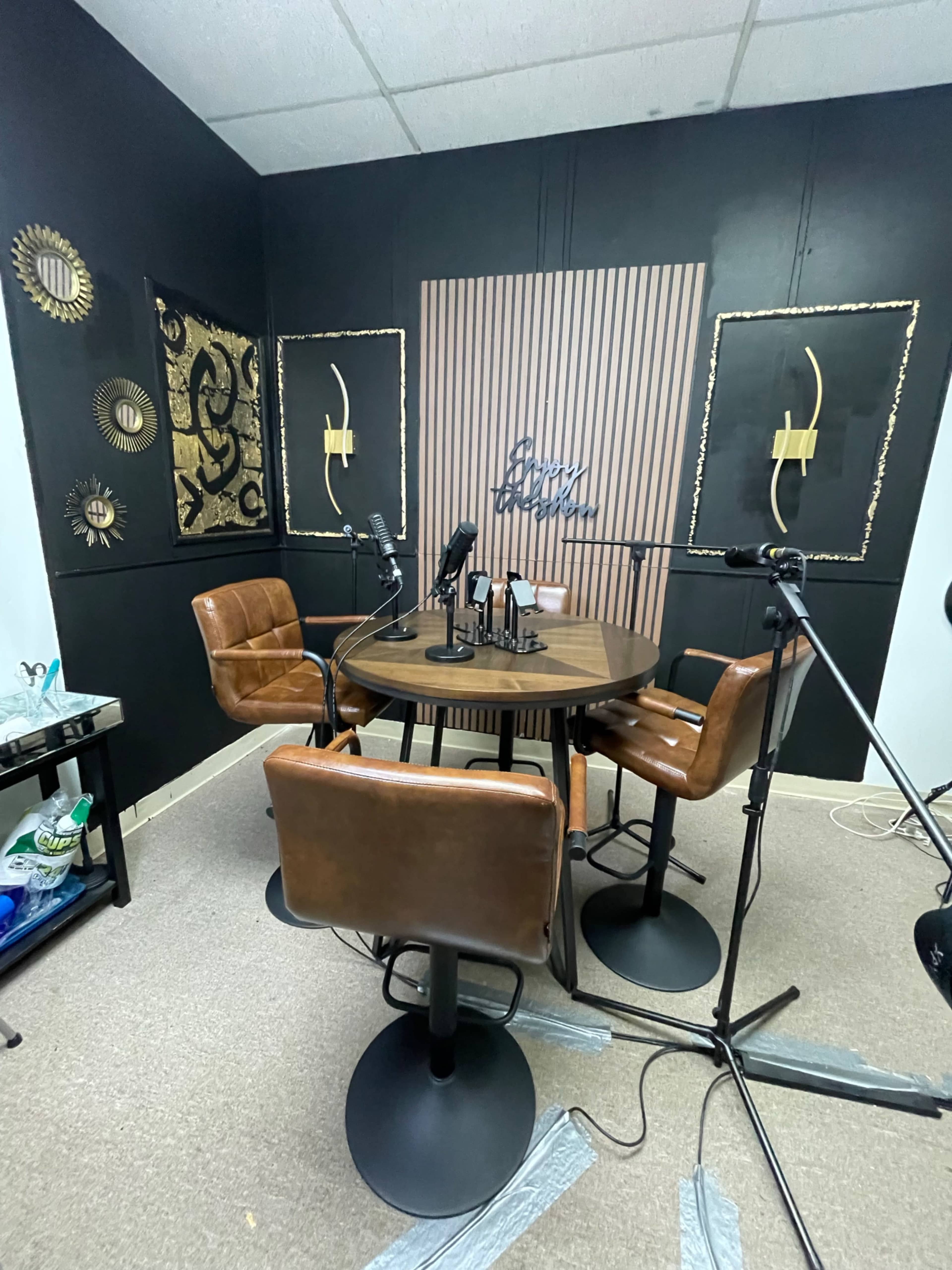 The image shows a podcast studio setup with a round table, four brown leather chairs, and multiple microphones positioned around the table against a black wall.
