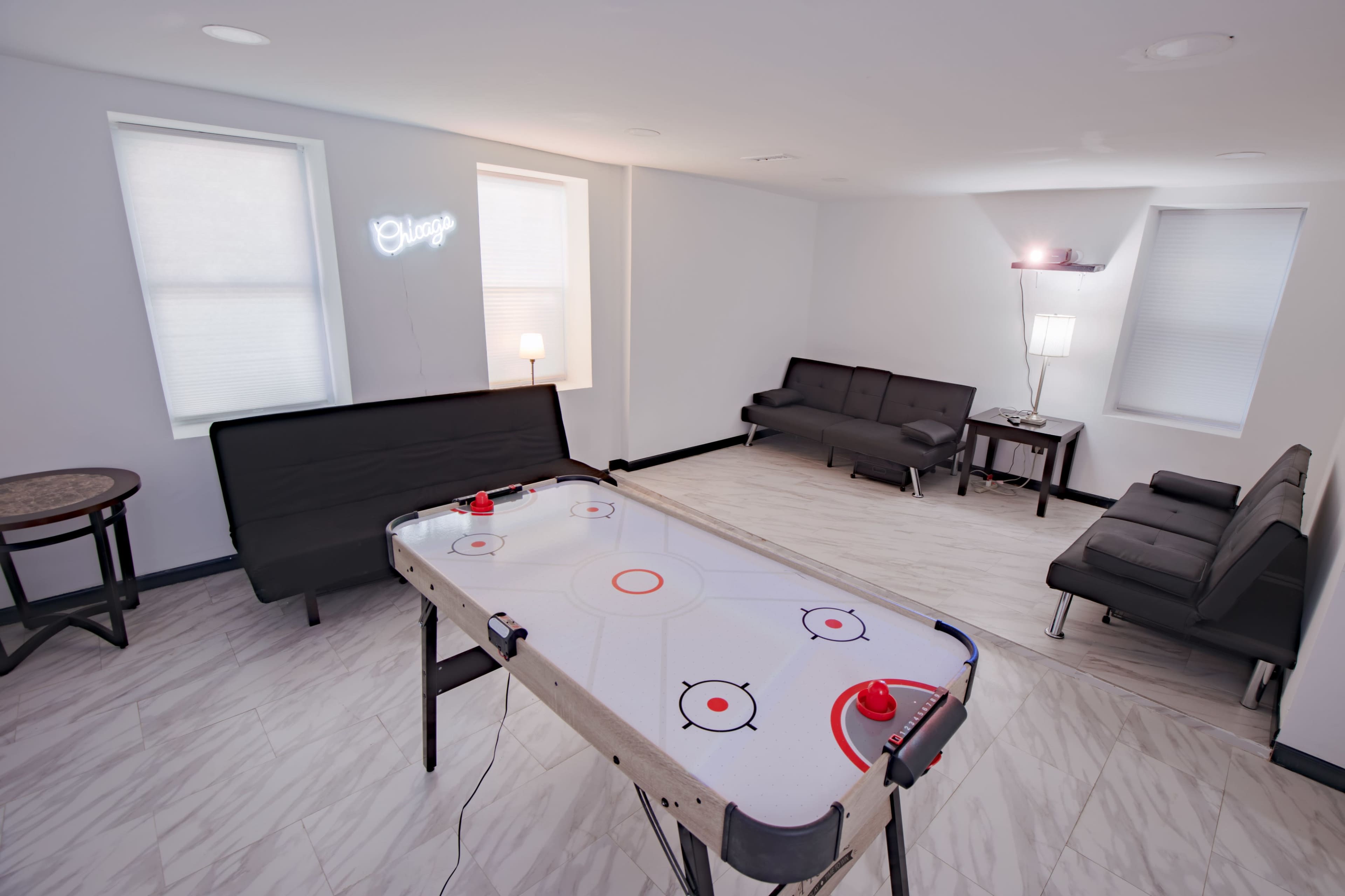 A modern, minimally furnished room featuring an air hockey table in the center, surrounded by seating areas with couches and a lamp.