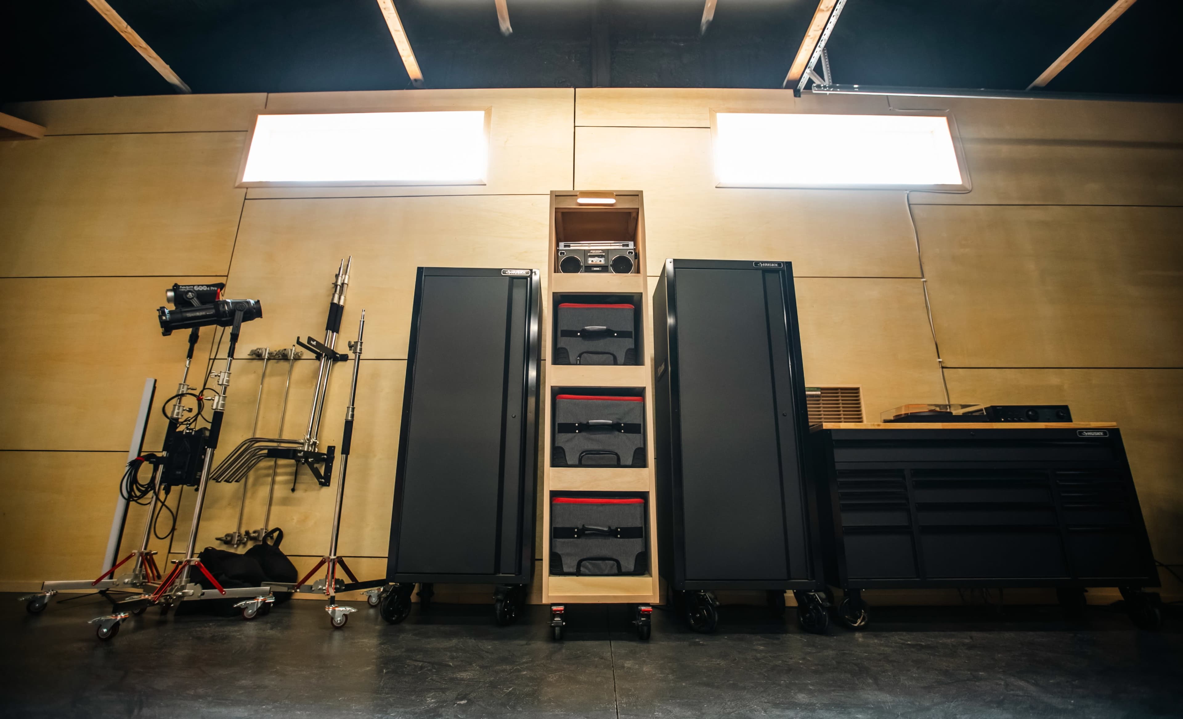 The image shows a modern workspace with two tall black cabinets, a stack of drawers in the center, and various equipment stands on the sides against a wooden wall.