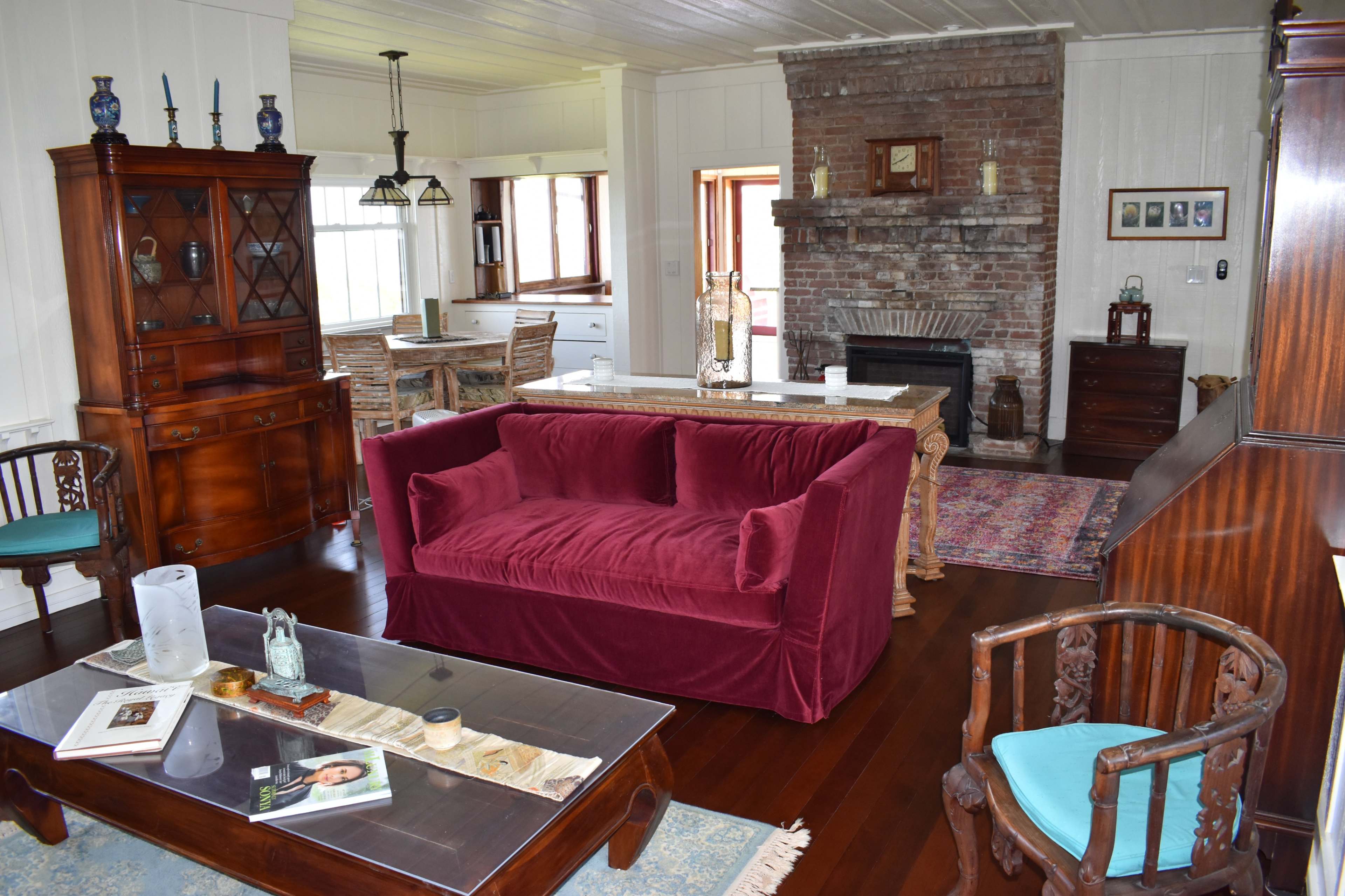 The image shows a cozy living room with a burgundy sofa, wooden furniture, a brick fireplace, and various decorative items arranged thoughtfully.