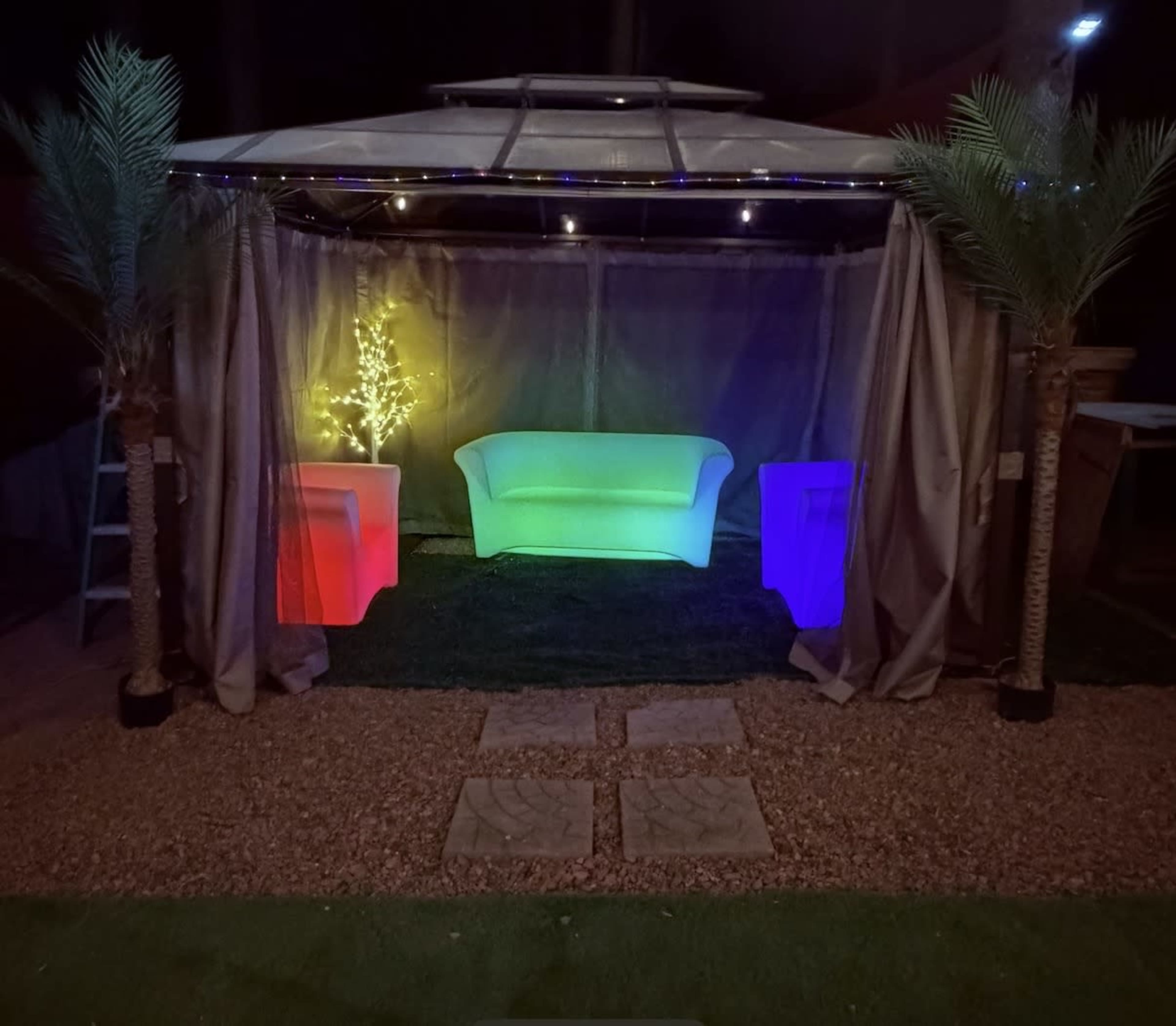 The scene features a colorful illuminated setup with a white couch and two glowing side tables under a gazebo, surrounded by gravel and grass.
