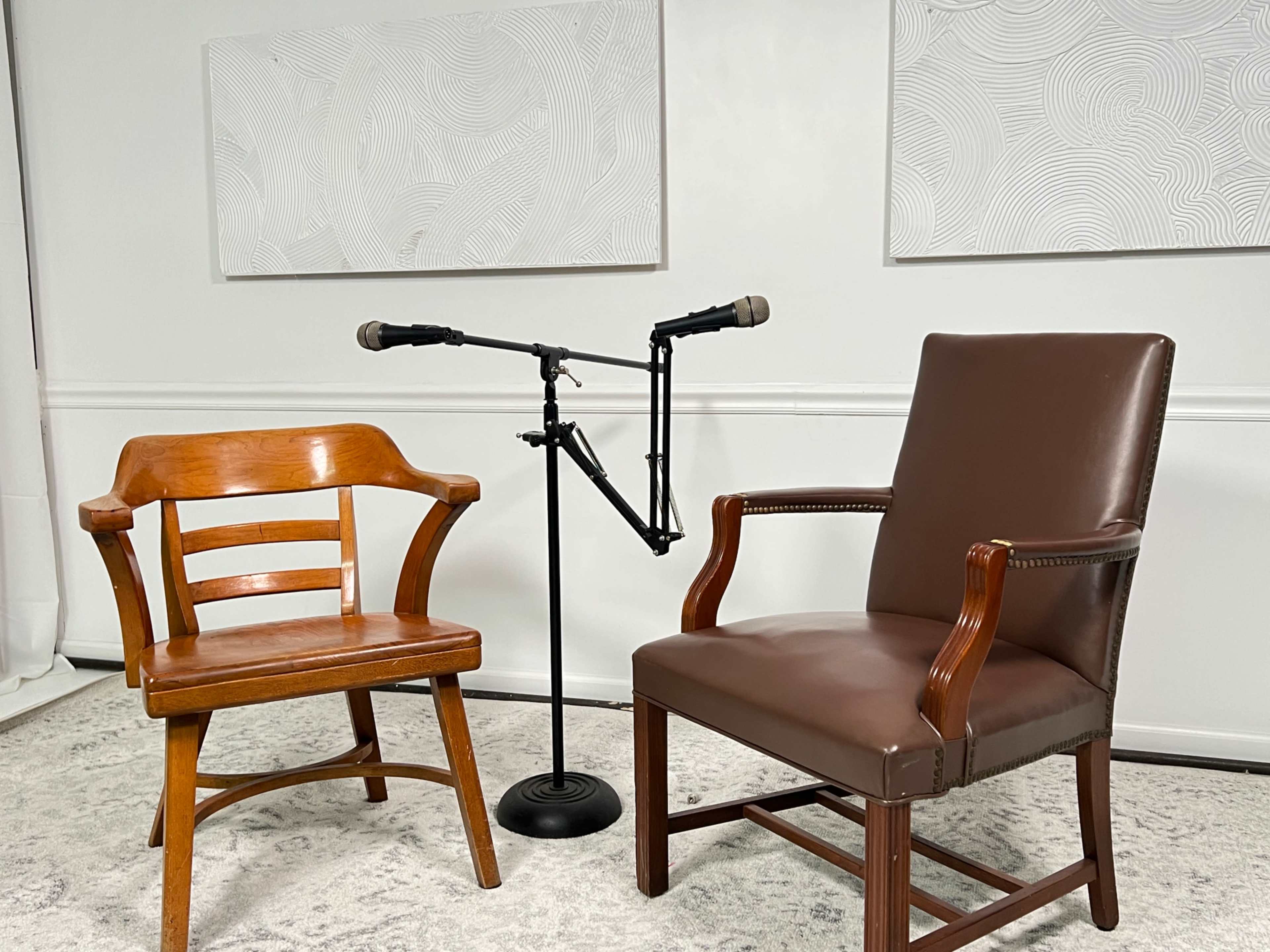 A wooden chair and a brown upholstered chair beside a microphone setup against a plain wall with abstract art.