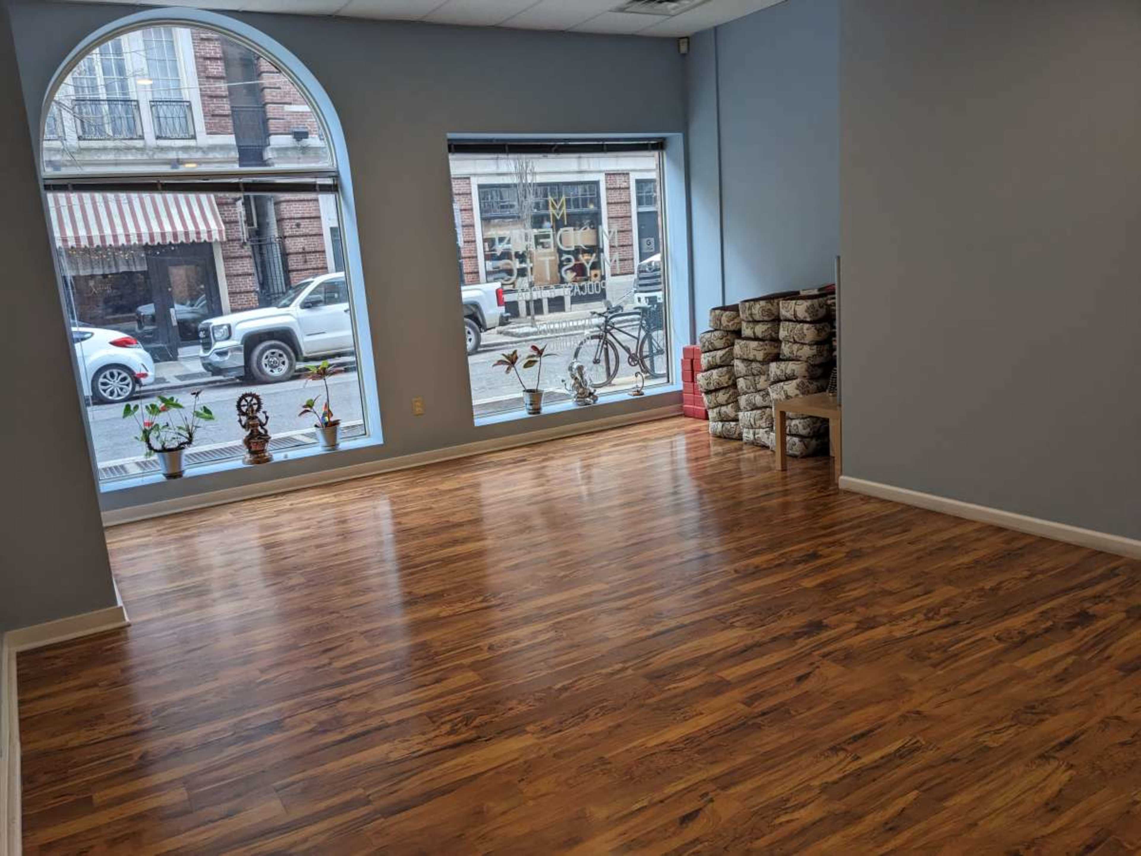 A spacious, empty room with wooden flooring and large windows displaying a street view.