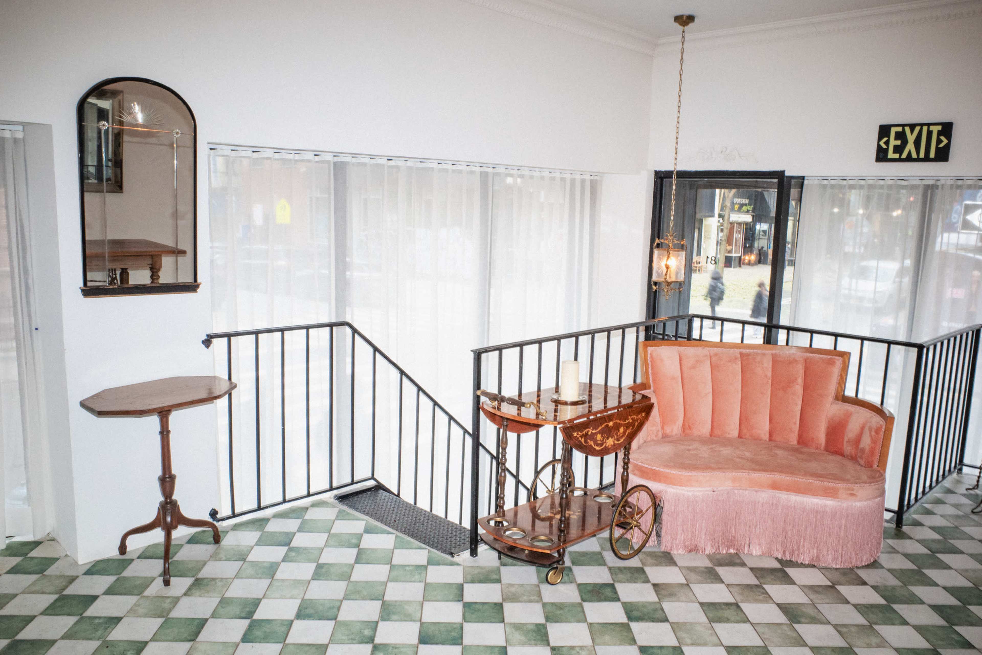 The image shows a seating area featuring a pink velvet couch with fringe, a small round table, and a mirror, all set against a patterned tile floor and a staircase railing.
