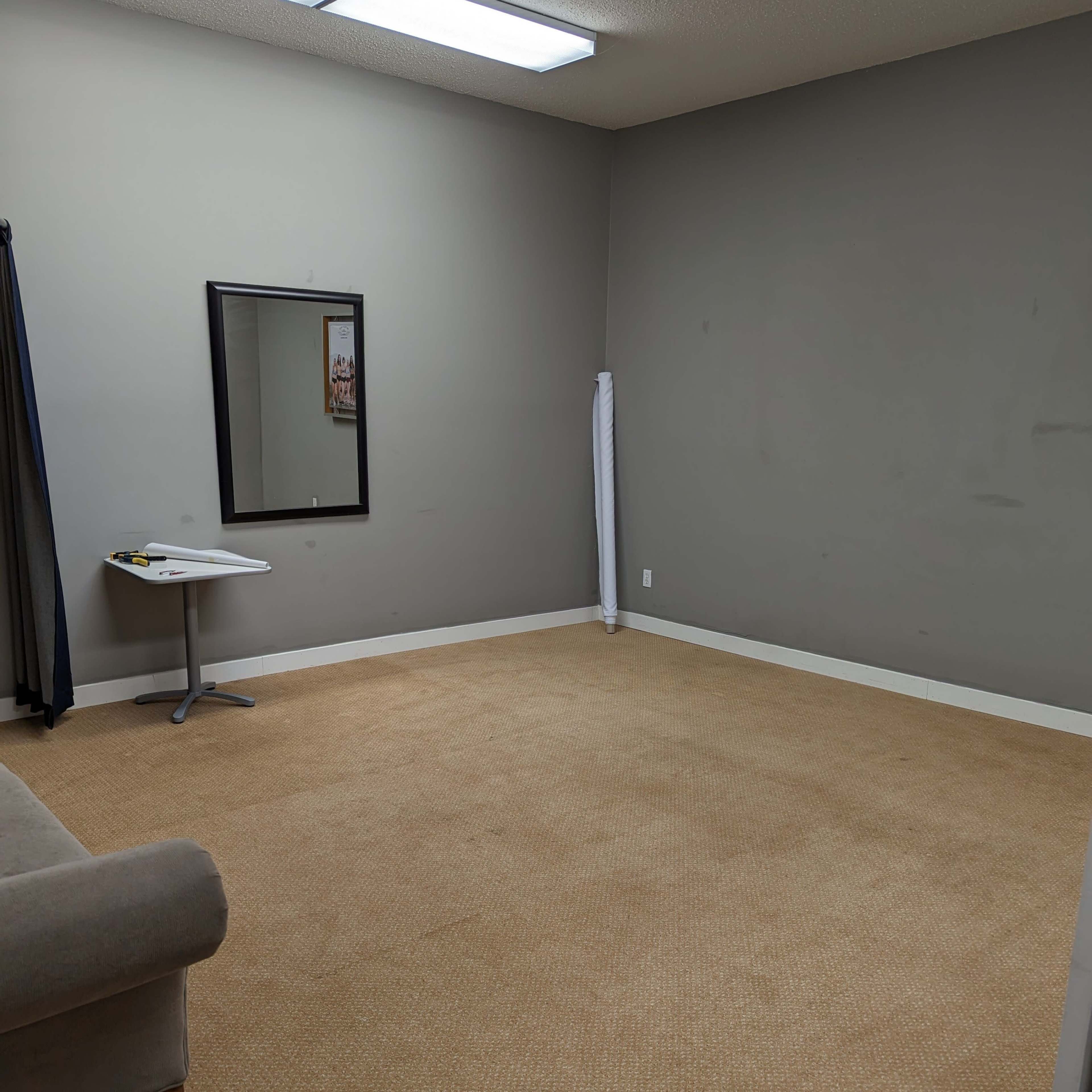A sparsely furnished room with gray walls, a tan carpet, a mirror on one wall, a small table, and a sofa in the corner.