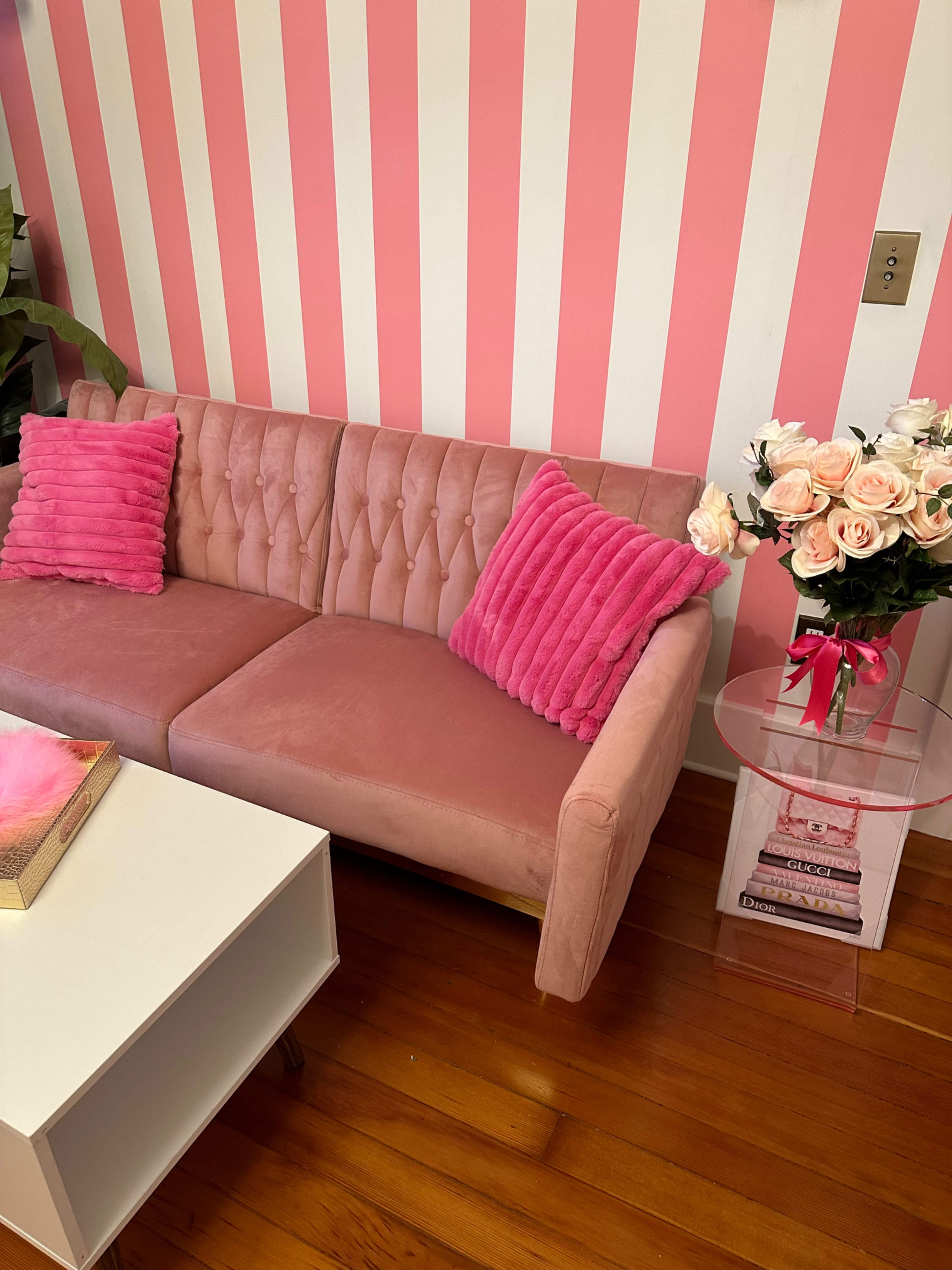 A pink tufted couch with matching cushions sits beside a clear side table adorned with a vase of roses and a small stack of books, against a striped pink wall.
