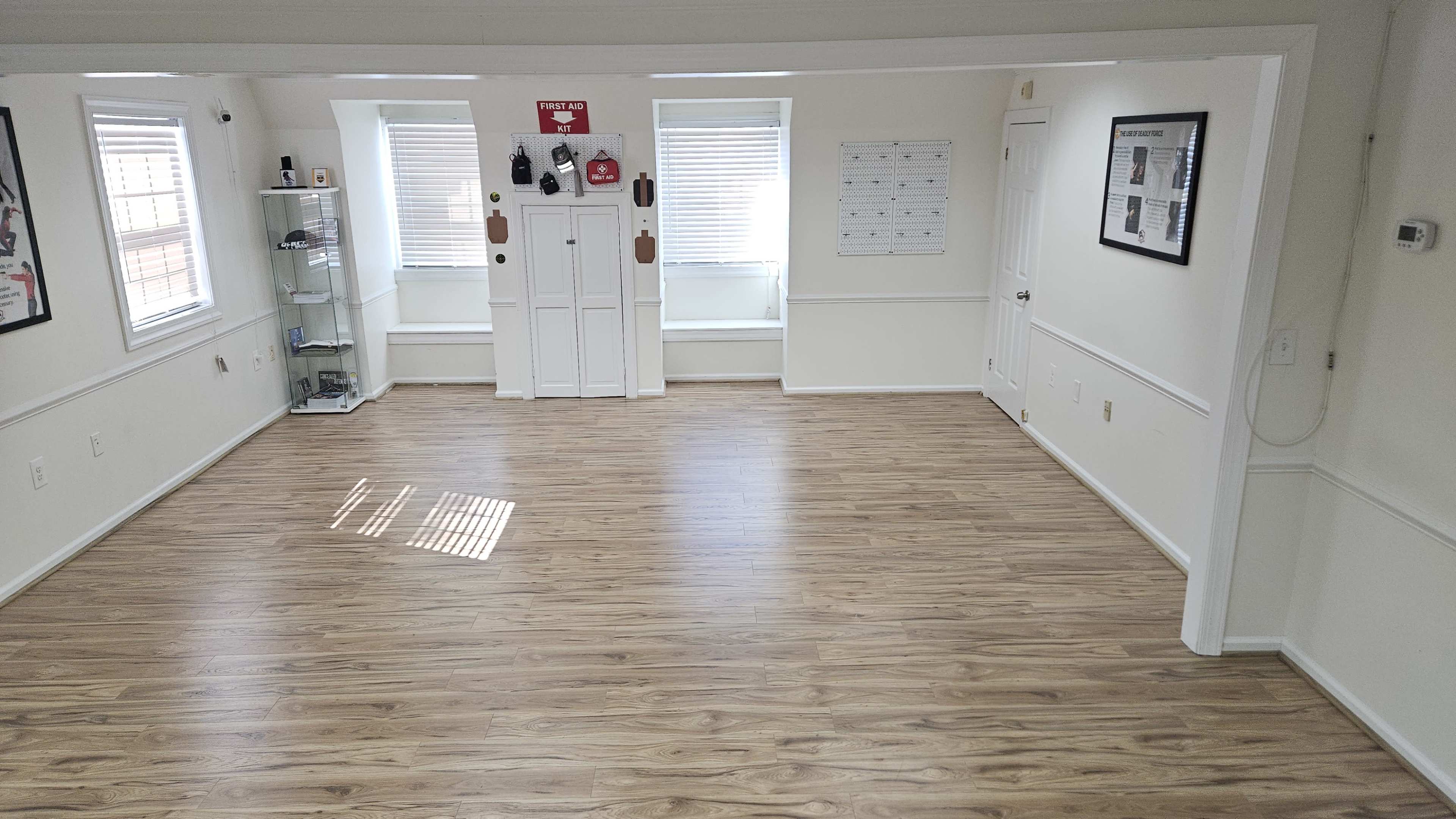 The image shows a spacious, empty room with light wooden flooring, white walls, and several windows allowing natural light.