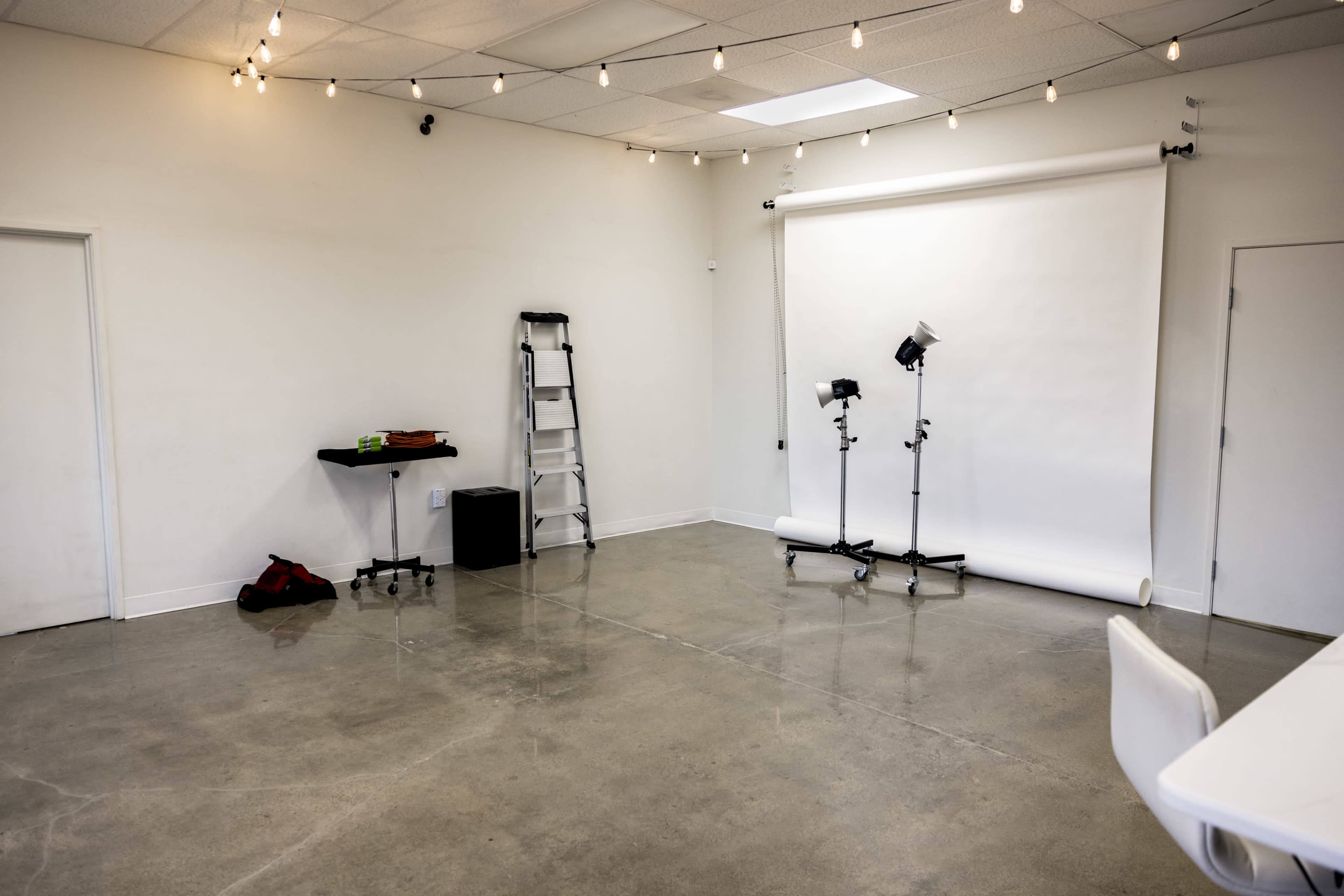 The image shows a spacious, well-lit studio with a white backdrop, a ladder, two light stands with softboxes, and minimal furniture.