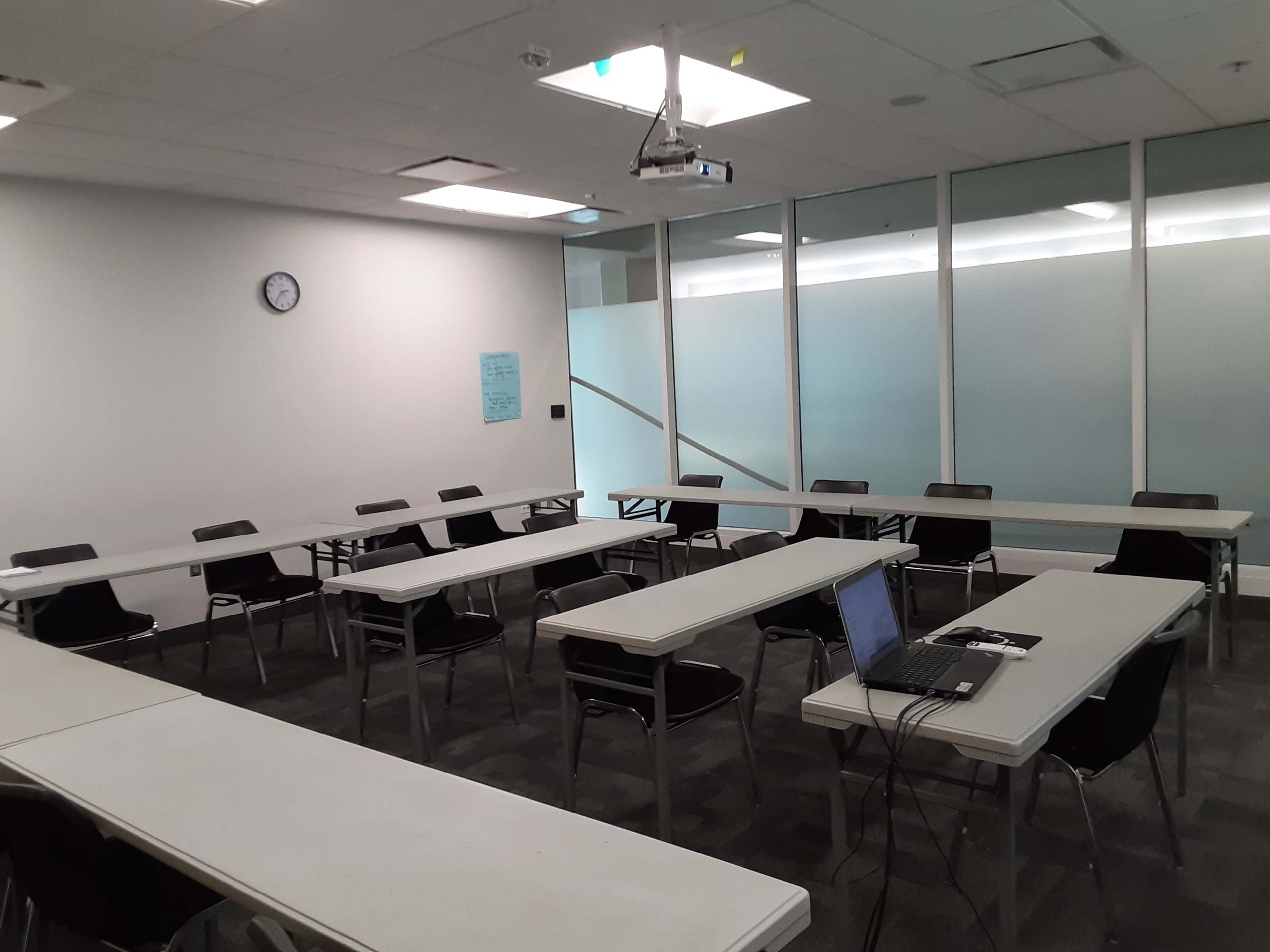 A modern classroom is set up with multiple white tables and black chairs, featuring a clock on the wall and a projector mounted on the ceiling.