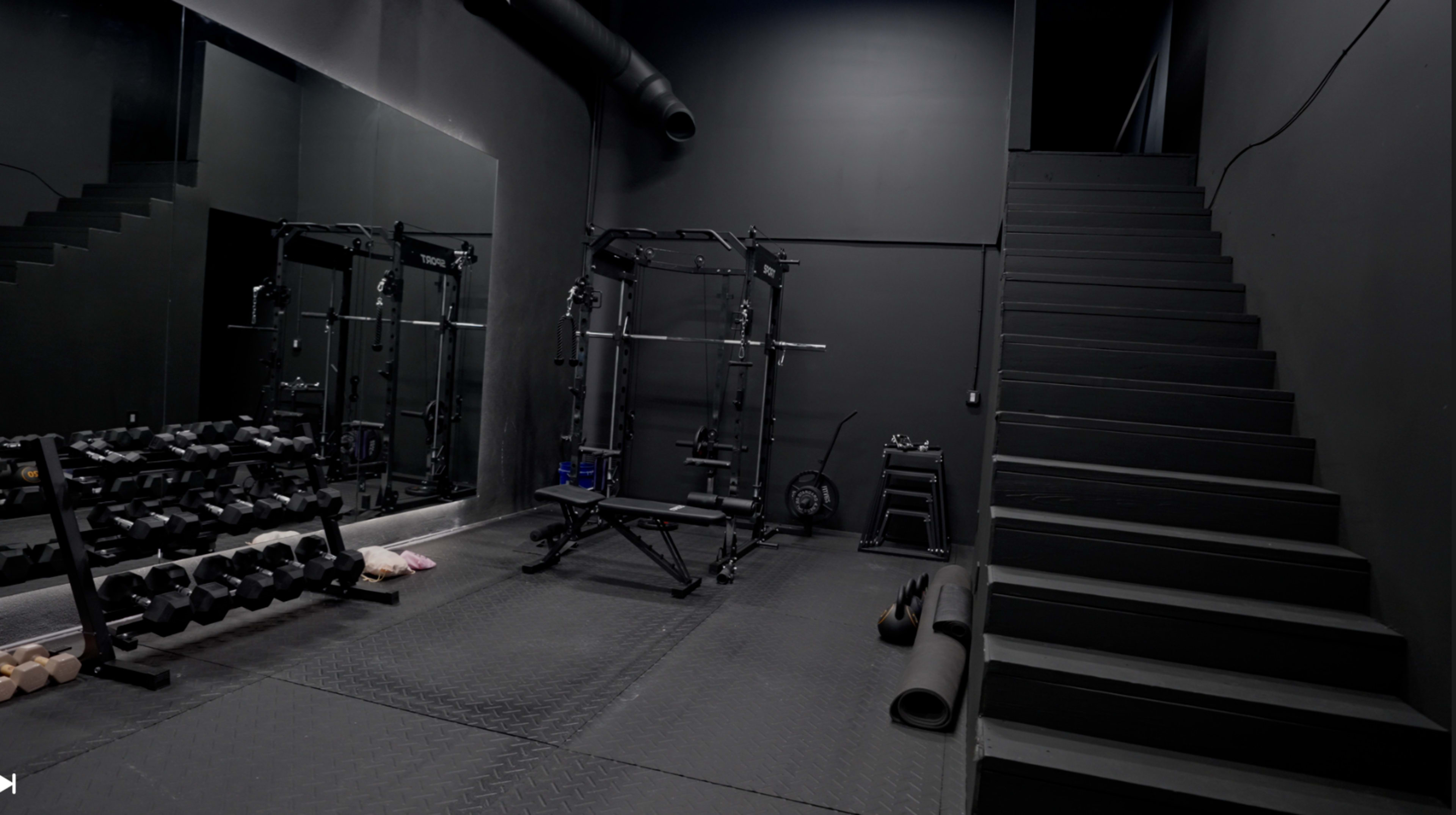 The image shows a dimly lit gym with a black interior, featuring weightlifting equipment, dumbbells, and a staircase leading to an upper level.