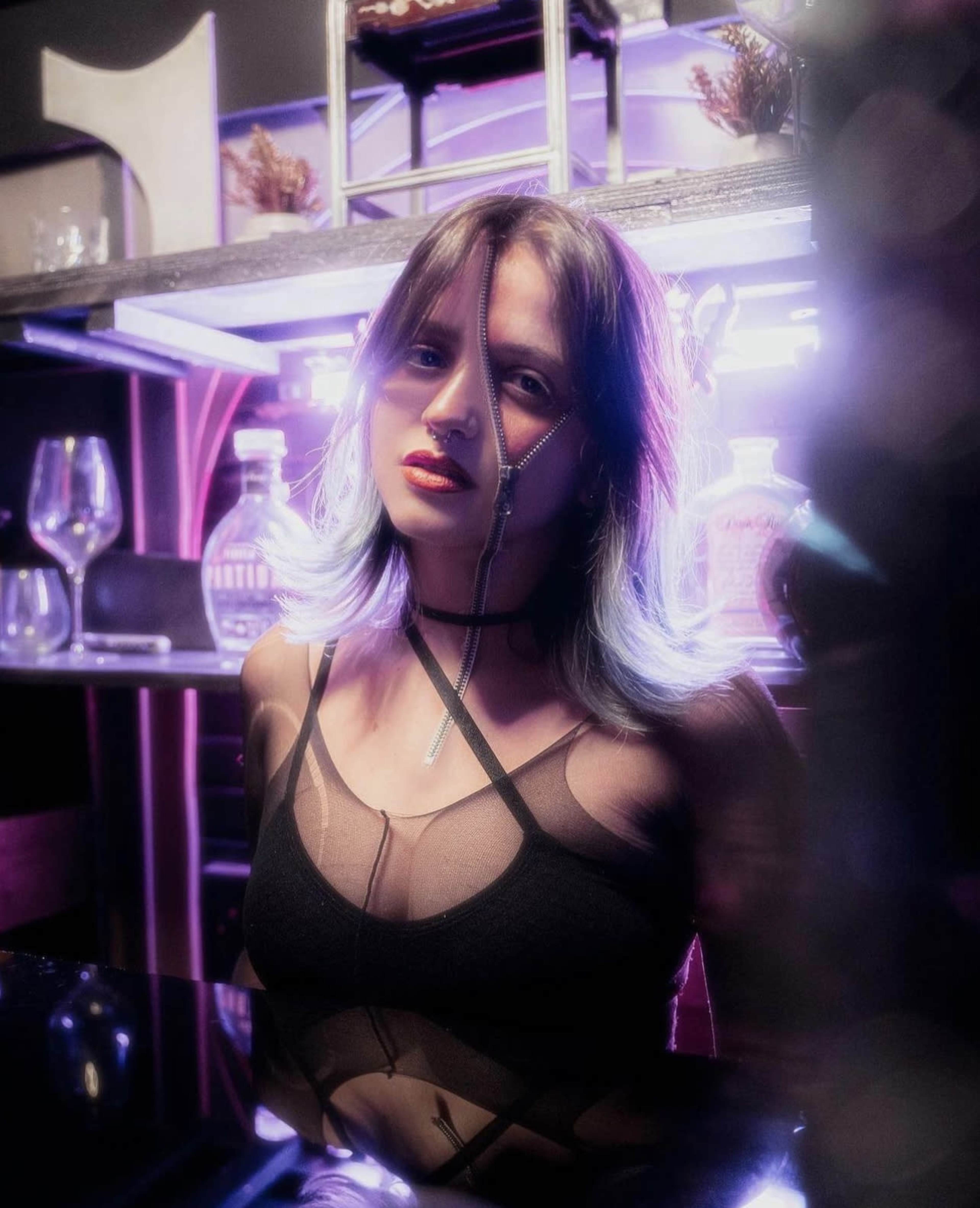 A woman with purple-tinted hair is standing in front of a bar with illuminated shelves and glassware.