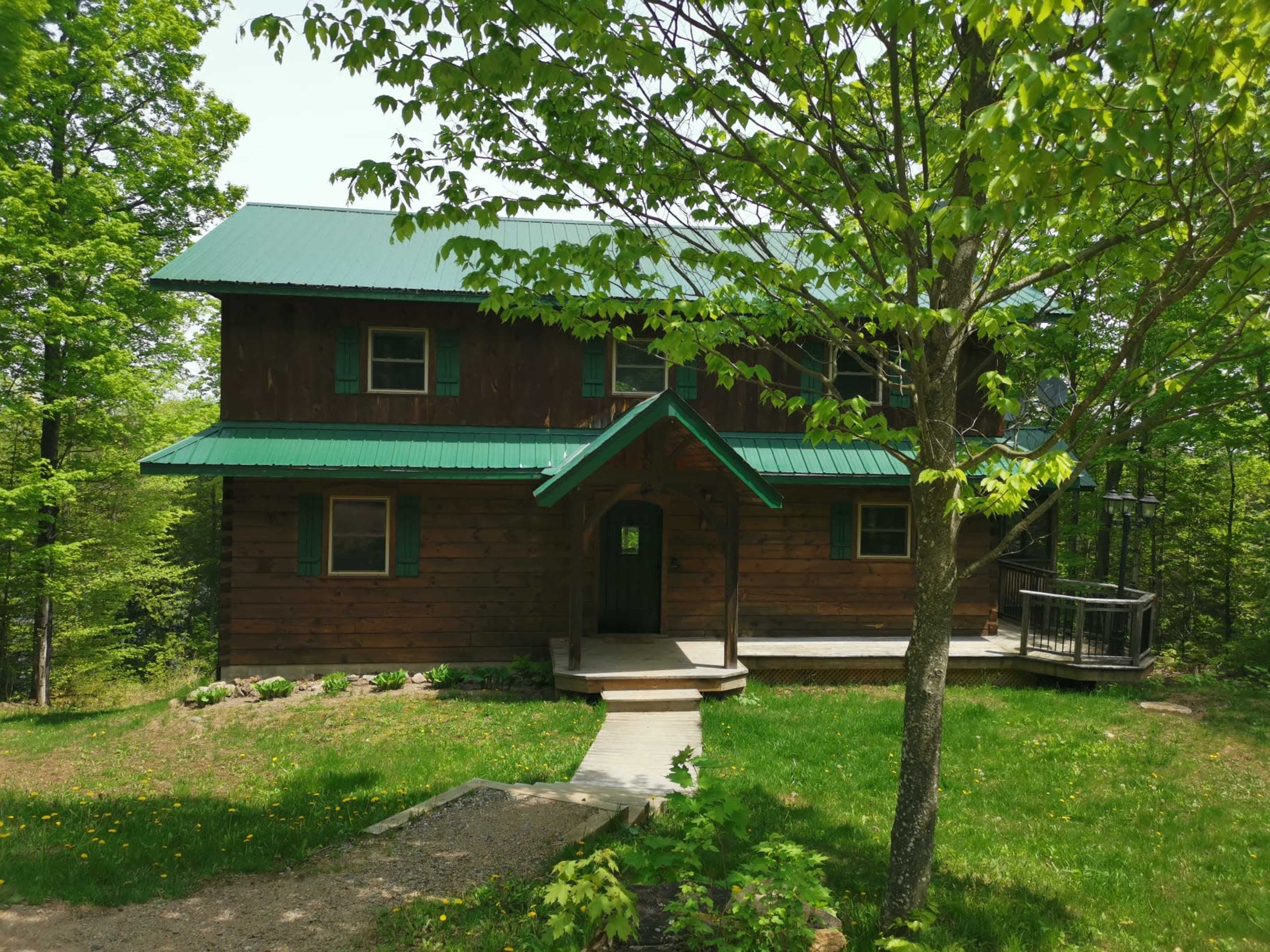 A two-story wooden house with a green metal roof is surrounded by trees and grass, featuring a front porch and walkway.