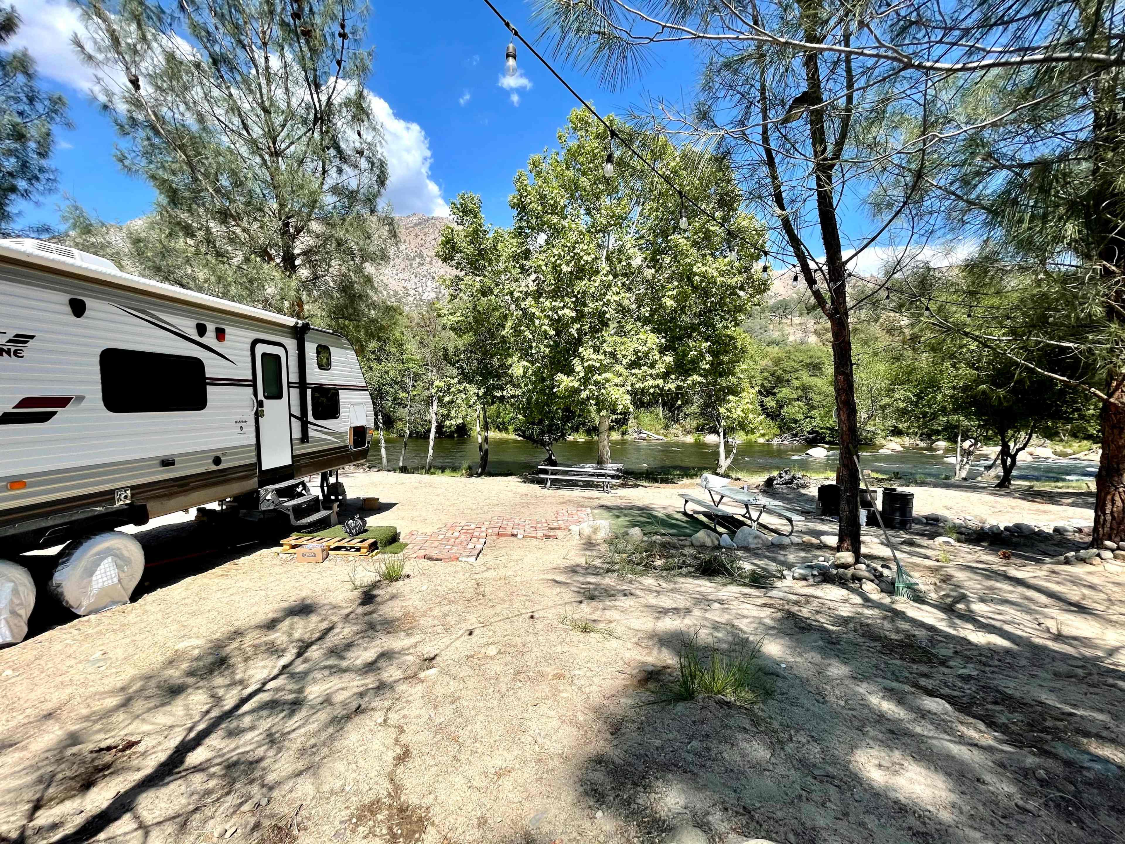A travel trailer is set up in a sandy area beside a river, surrounded by trees and outdoor seating.