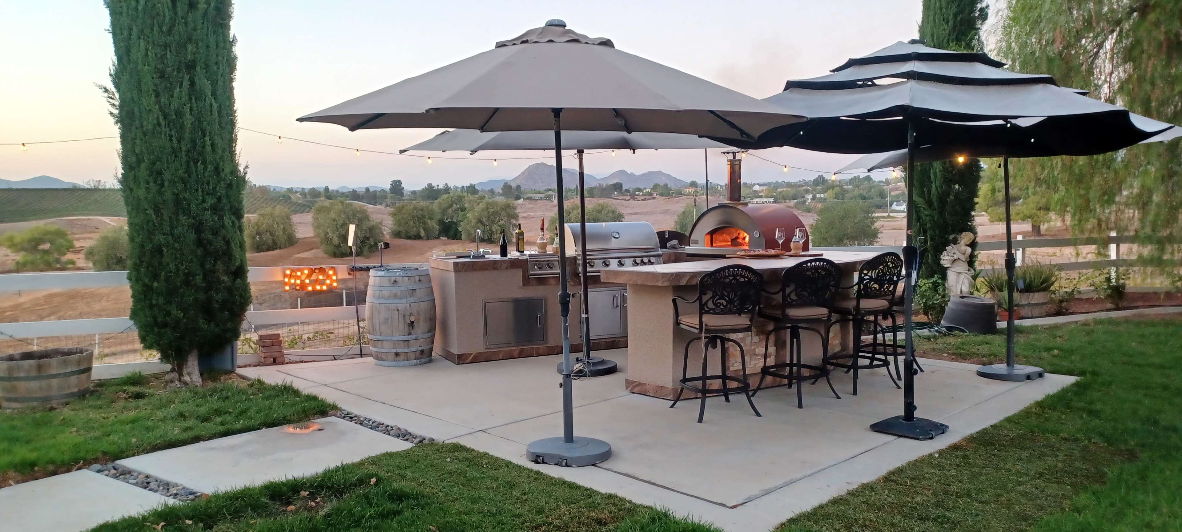 The image shows an outdoor kitchen area with a bar, two large umbrellas, and a pizza oven, surrounded by a grassy space and distant mountains.