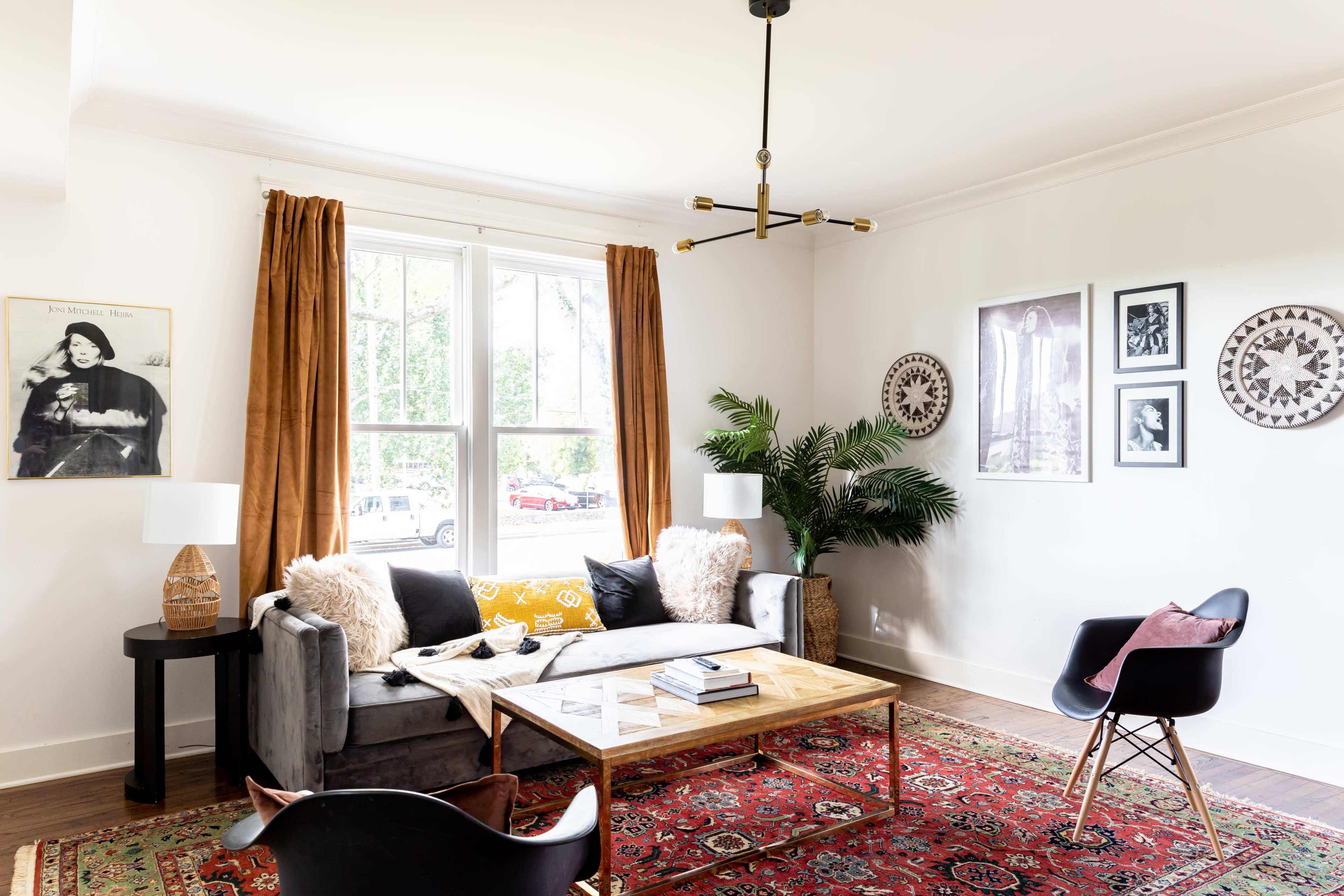 A living room features a gray sofa with decorative pillows, a wooden coffee table, and a mix of plants and artwork along the walls.