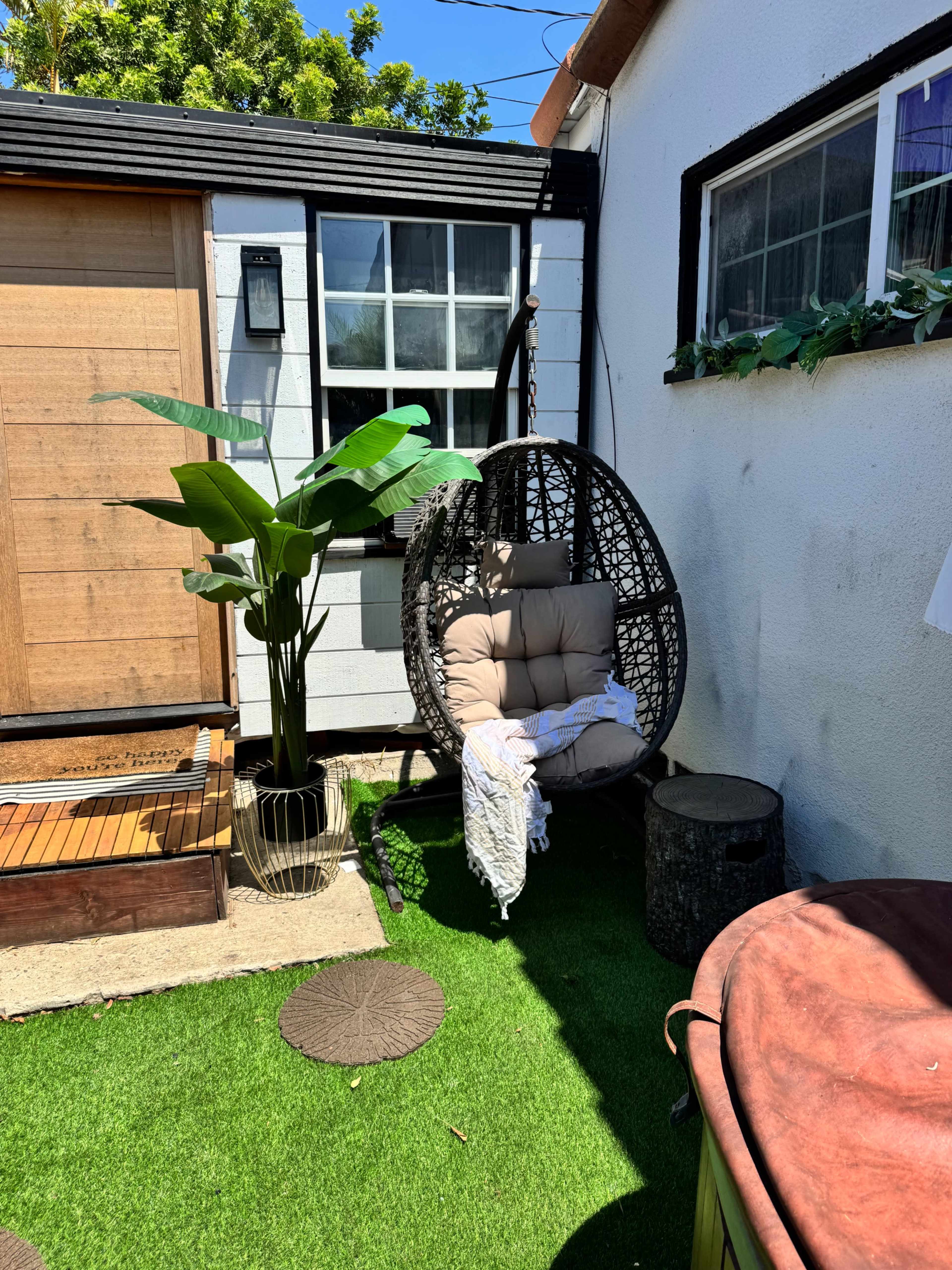 The image shows a cozy outdoor area featuring a hanging chair, a potted plant, and a wooden cabin with a large window.