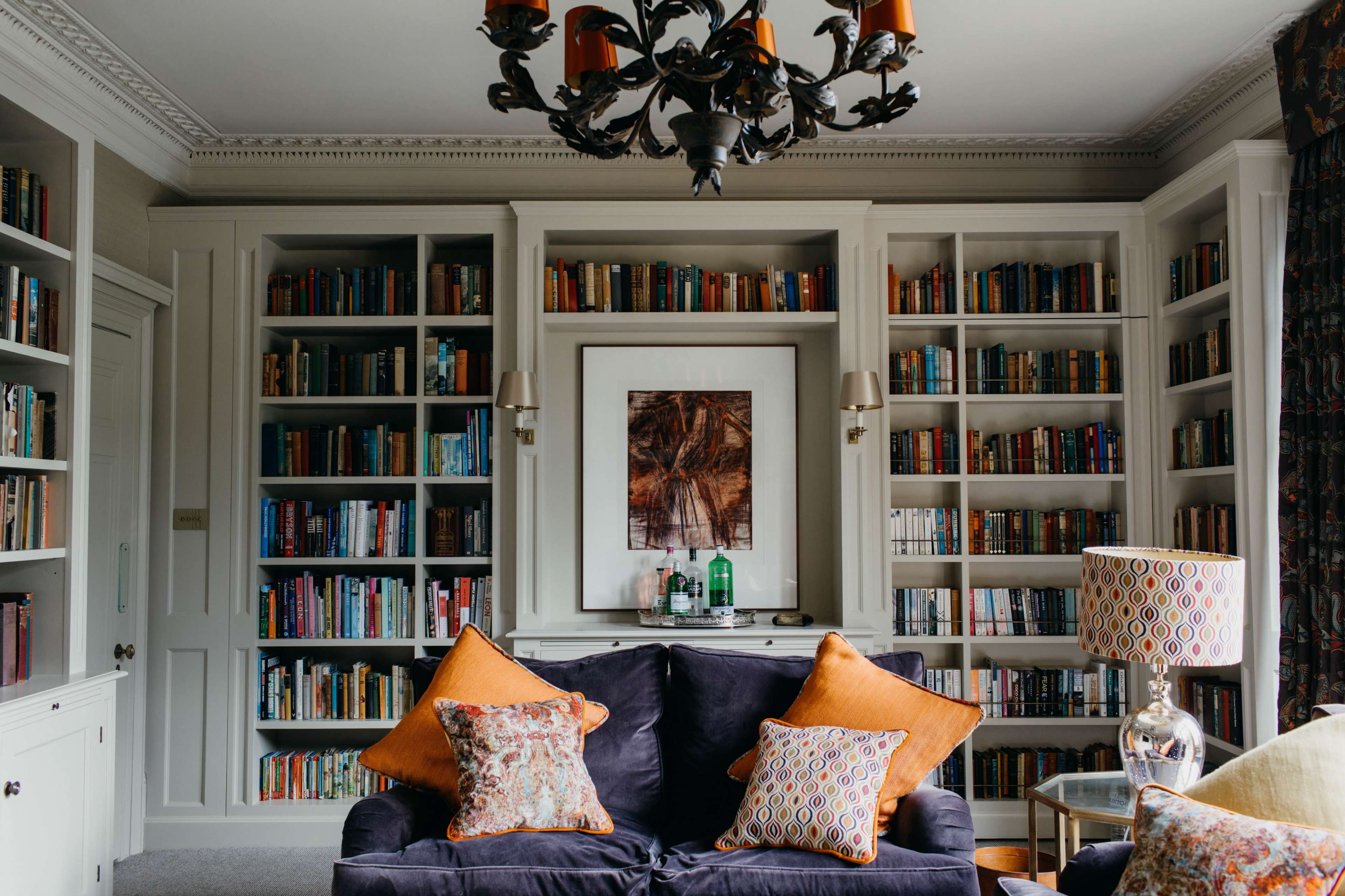 A cozy living room with a dark sofa, decorative pillows, a large bookshelf filled with books, and a chandelier overhead.