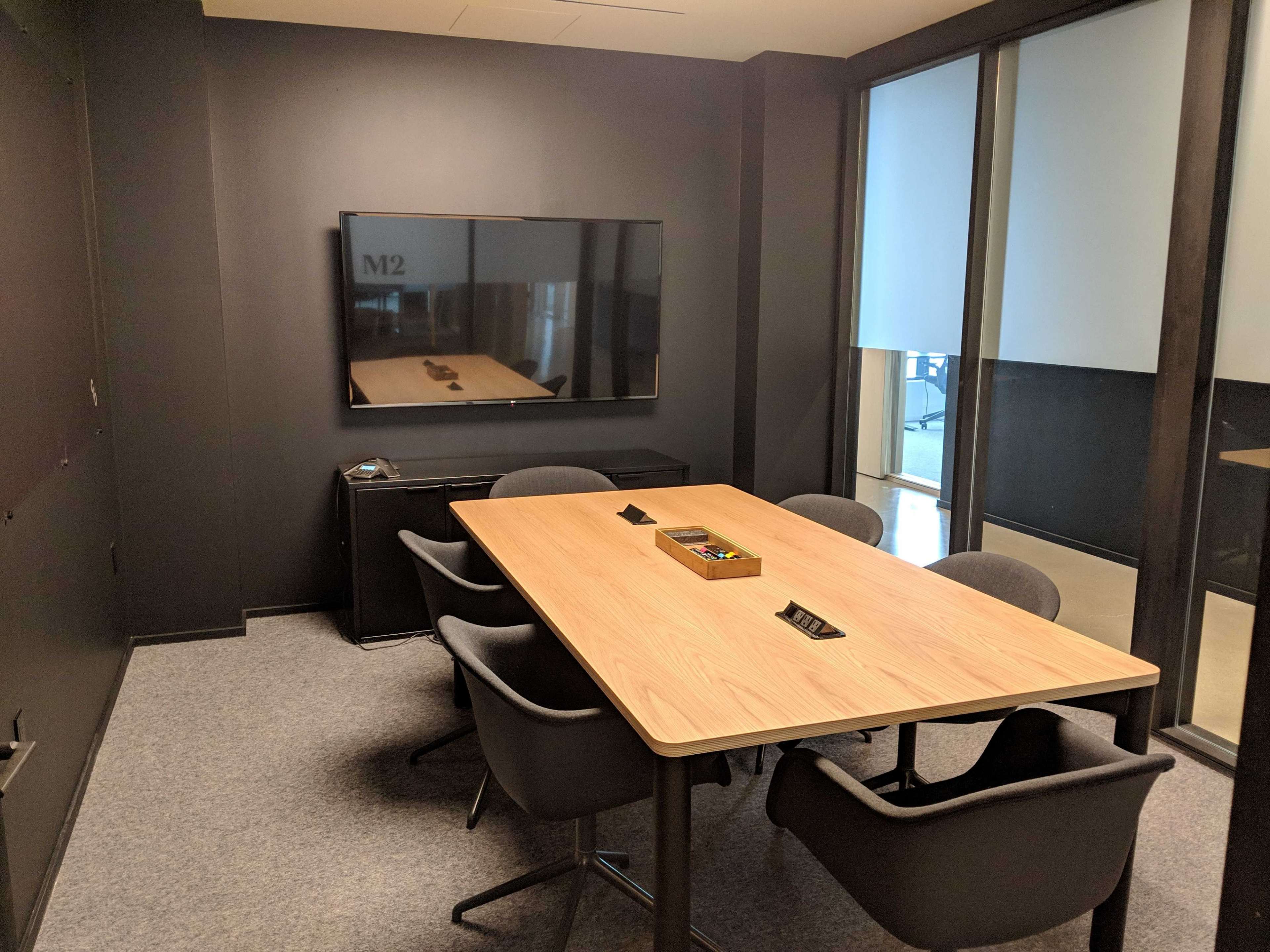 A conference room features a wooden table surrounded by black chairs, with a television mounted on the wall opposite a glass partition.