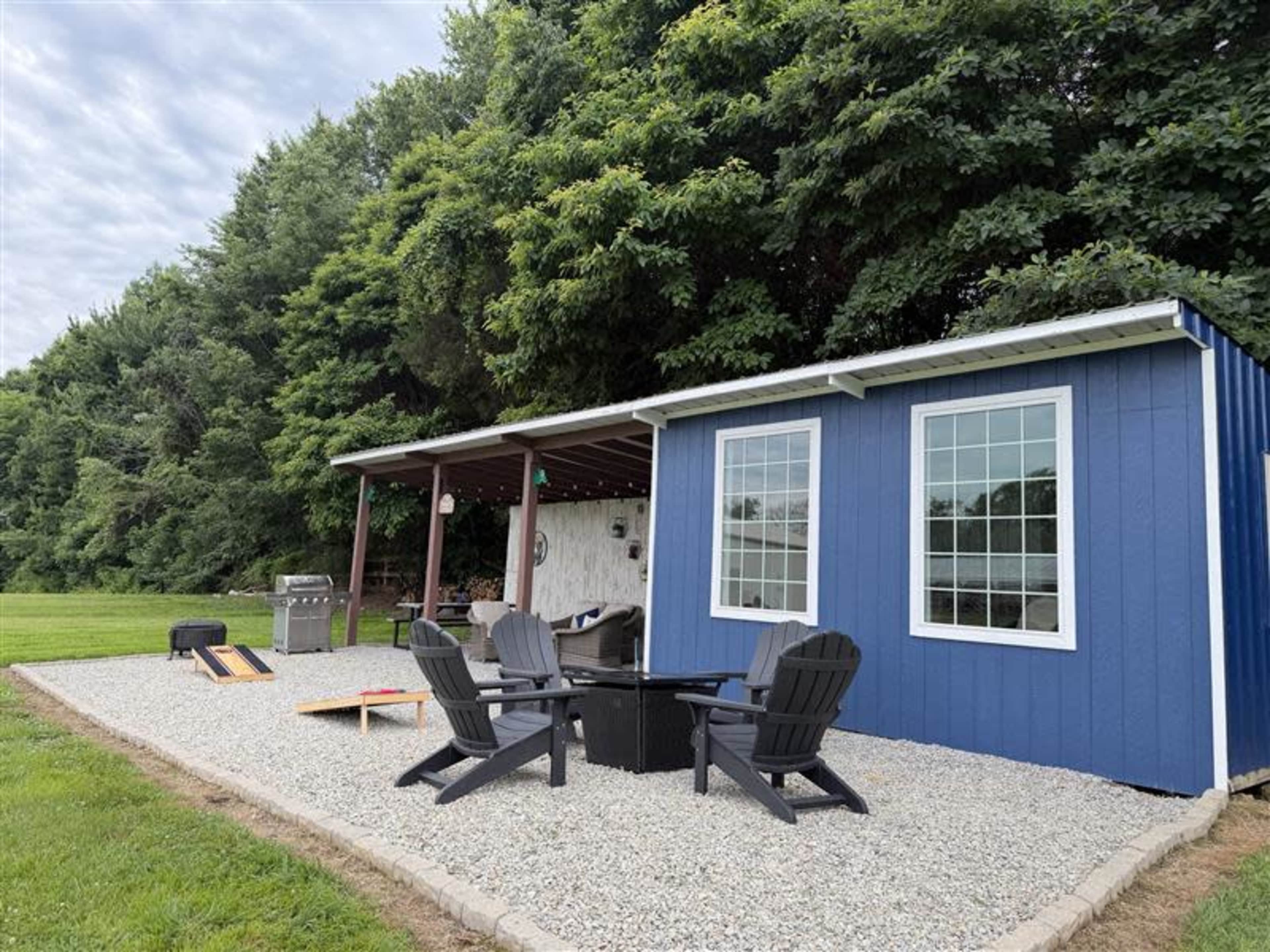 The image shows a small blue cabin with large windows, situated next to a covered porch area and surrounded by a gravel patio with seating and a grill.
