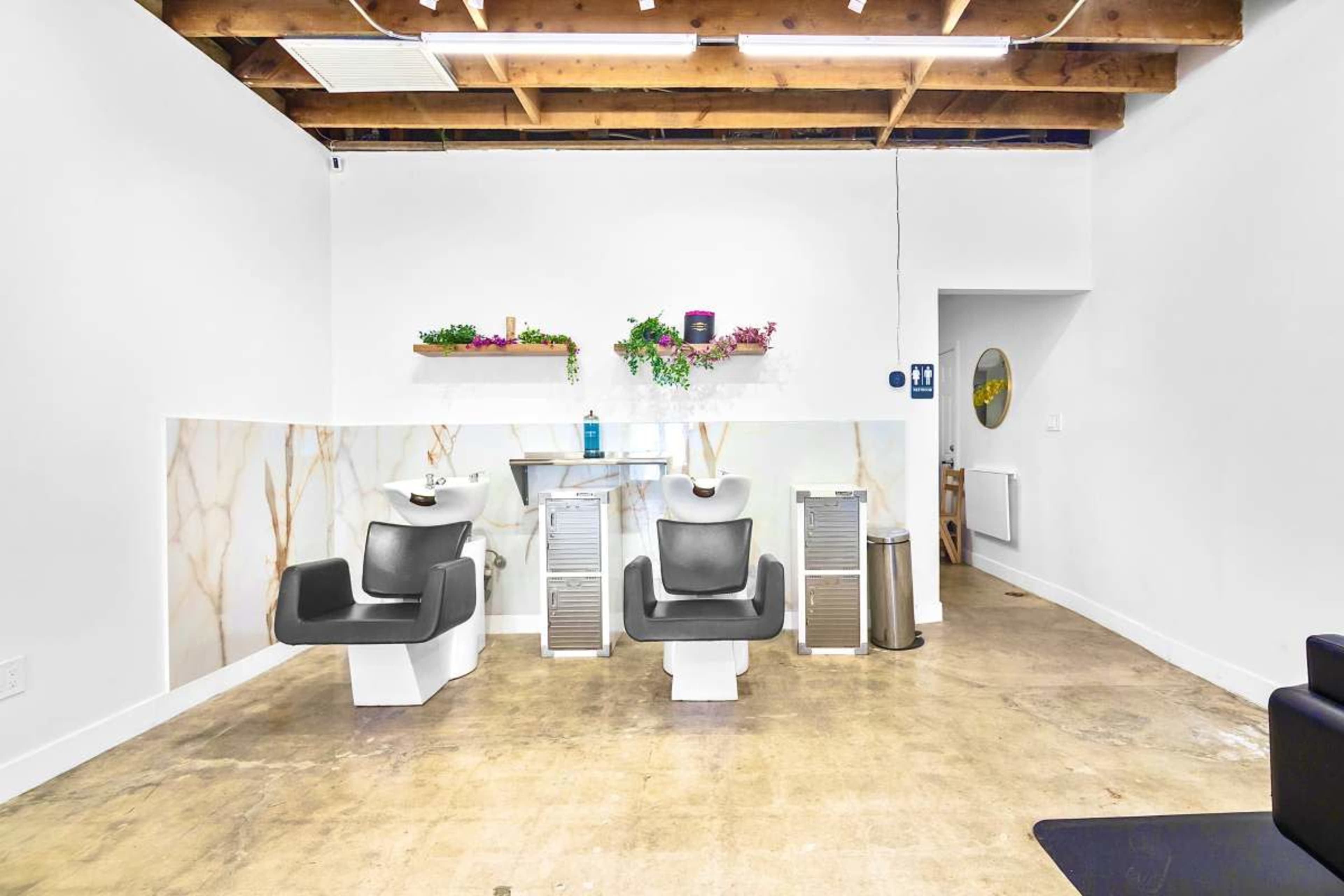 The image shows a salon interior featuring two styling chairs, a sink station, and minimalist decor against a white wall with a wooden ceiling.