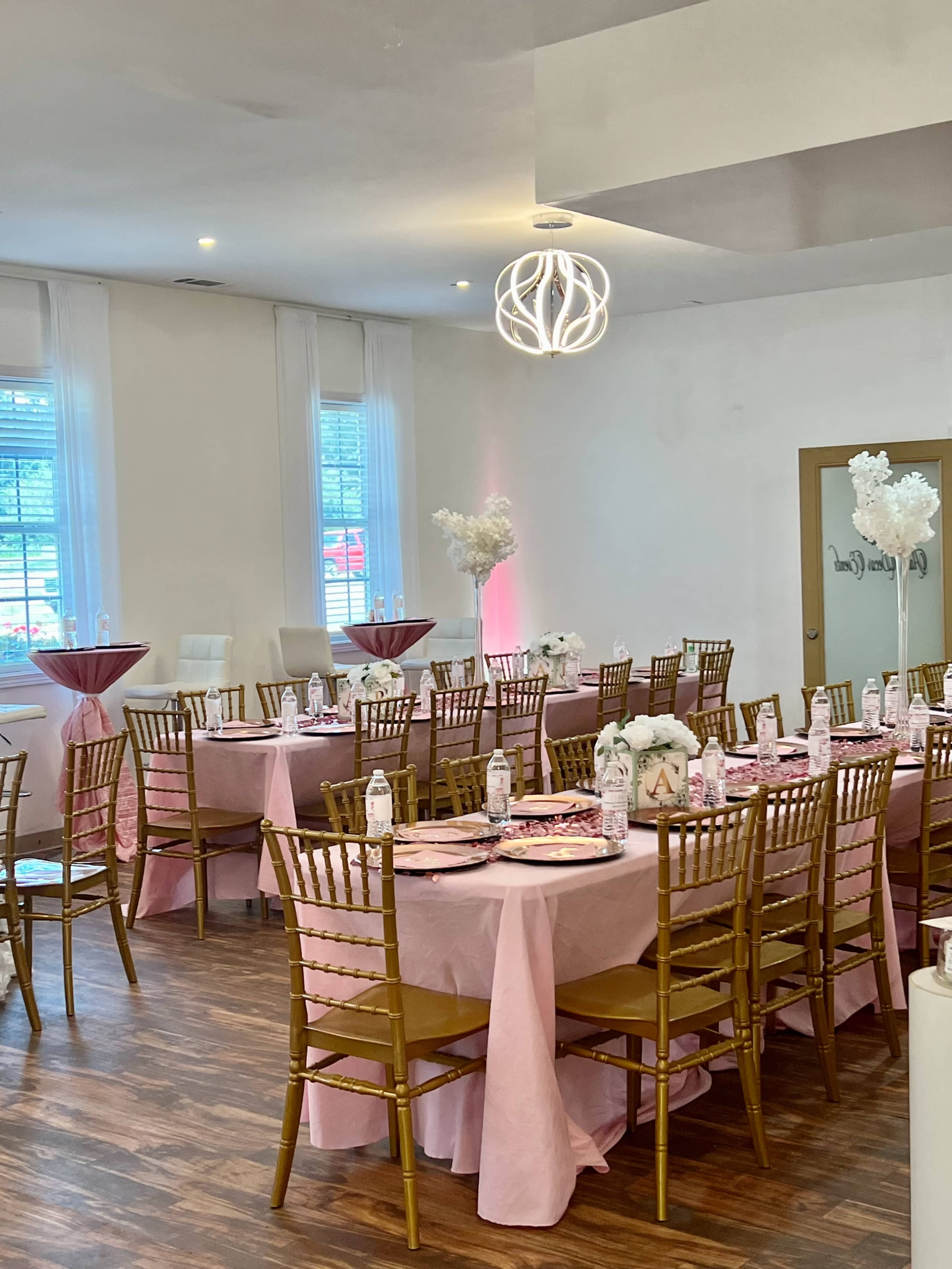 The image shows a decorated event space with tables covered in pink tablecloths, gold chiavari chairs, and floral centerpieces under modern lighting.