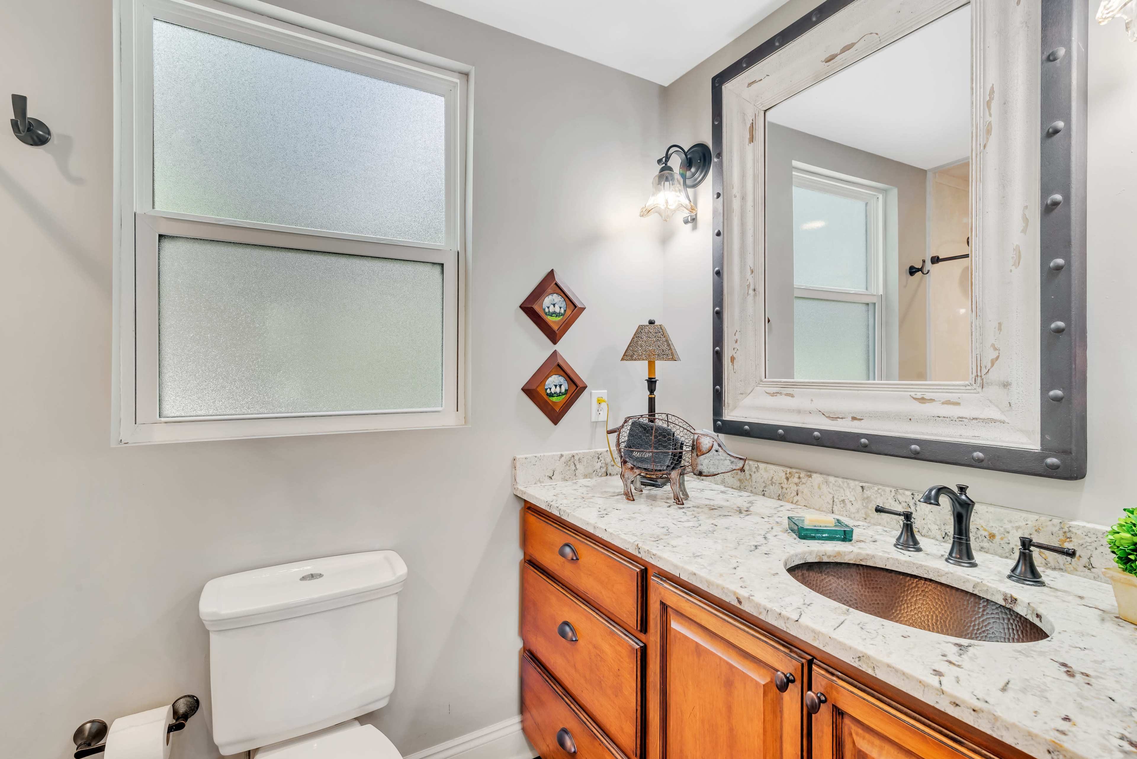 A bathroom featuring a toilet, a wooden vanity with a granite countertop, a mirror with a metal frame, and a window with frosted panes.