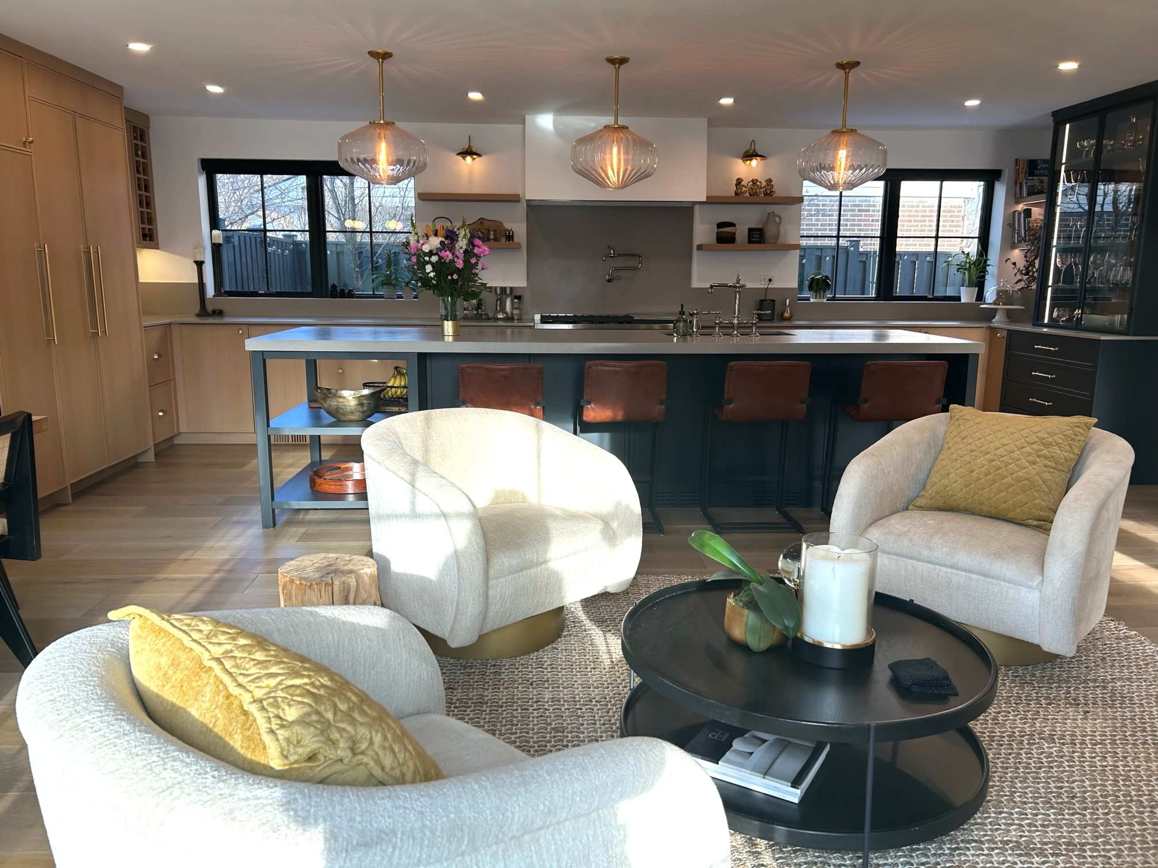 The image shows a modern kitchen and living area with three upholstered armchairs, a round coffee table, and an island with bar stools, all illuminated by pendant lights.