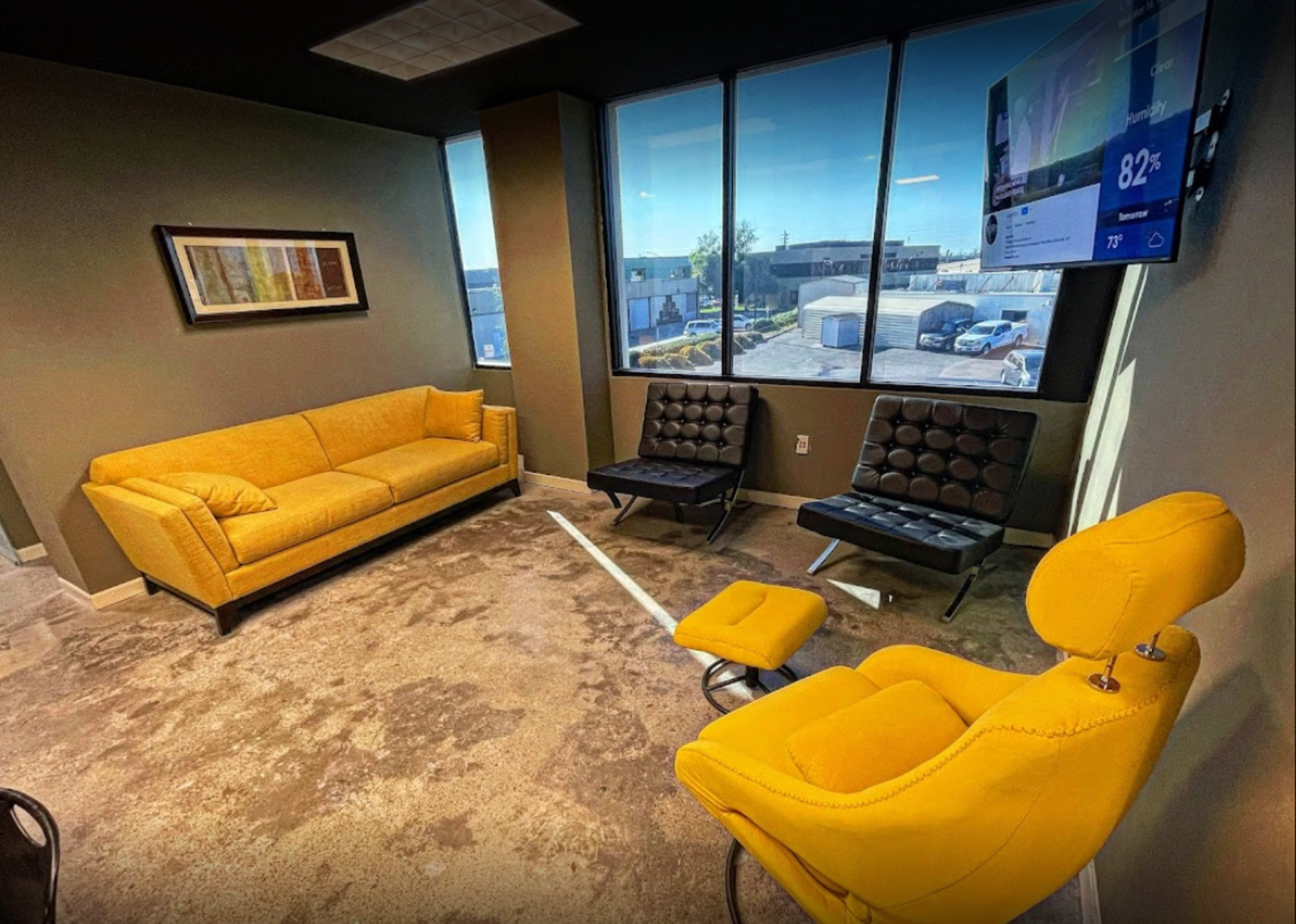 A modern office lounge with yellow furniture, including a sofa and two chairs, positioned around a carpeted floor and large windows.