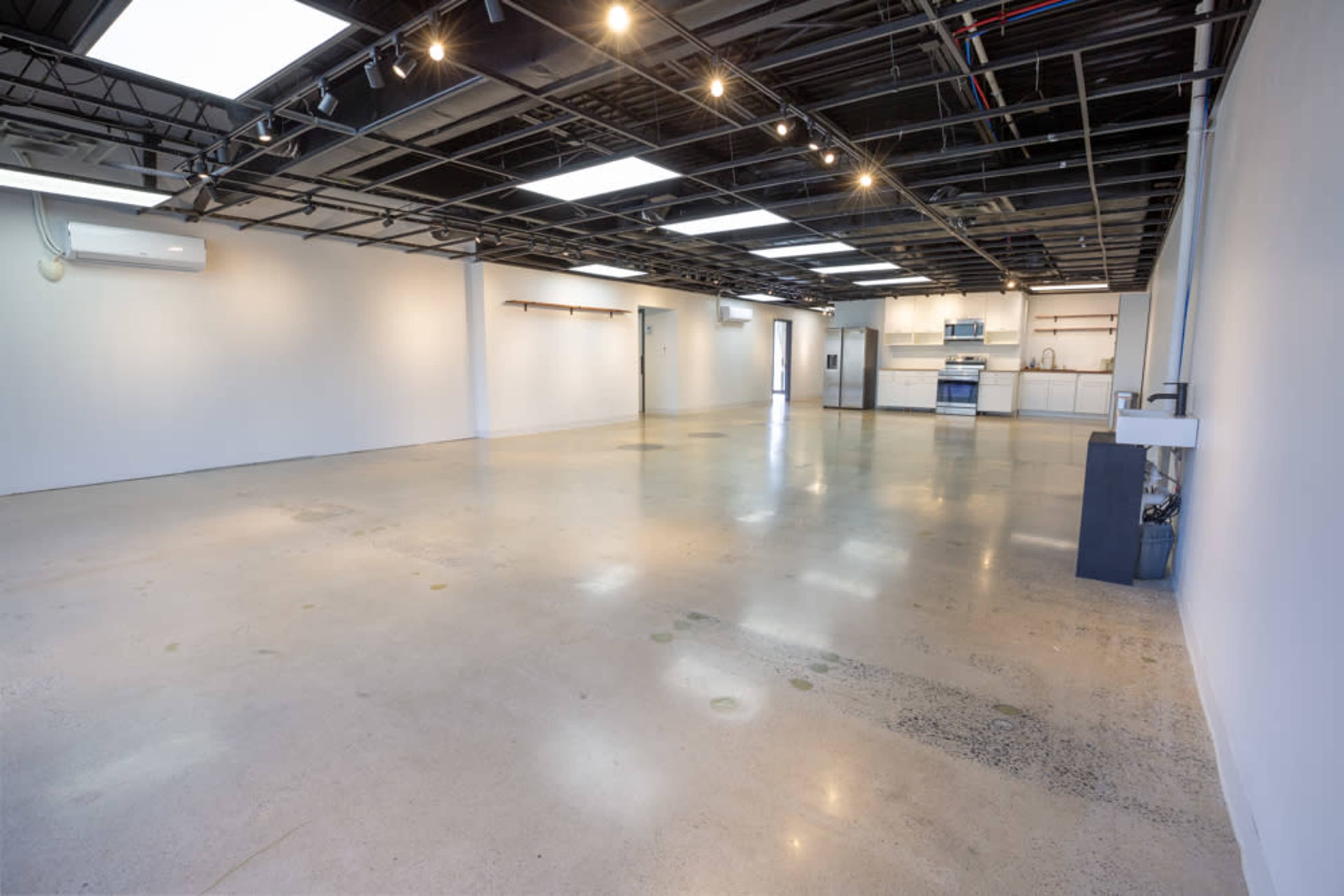 The image shows a spacious, empty interior room with polished concrete floors and a partially visible kitchen area at the far end.