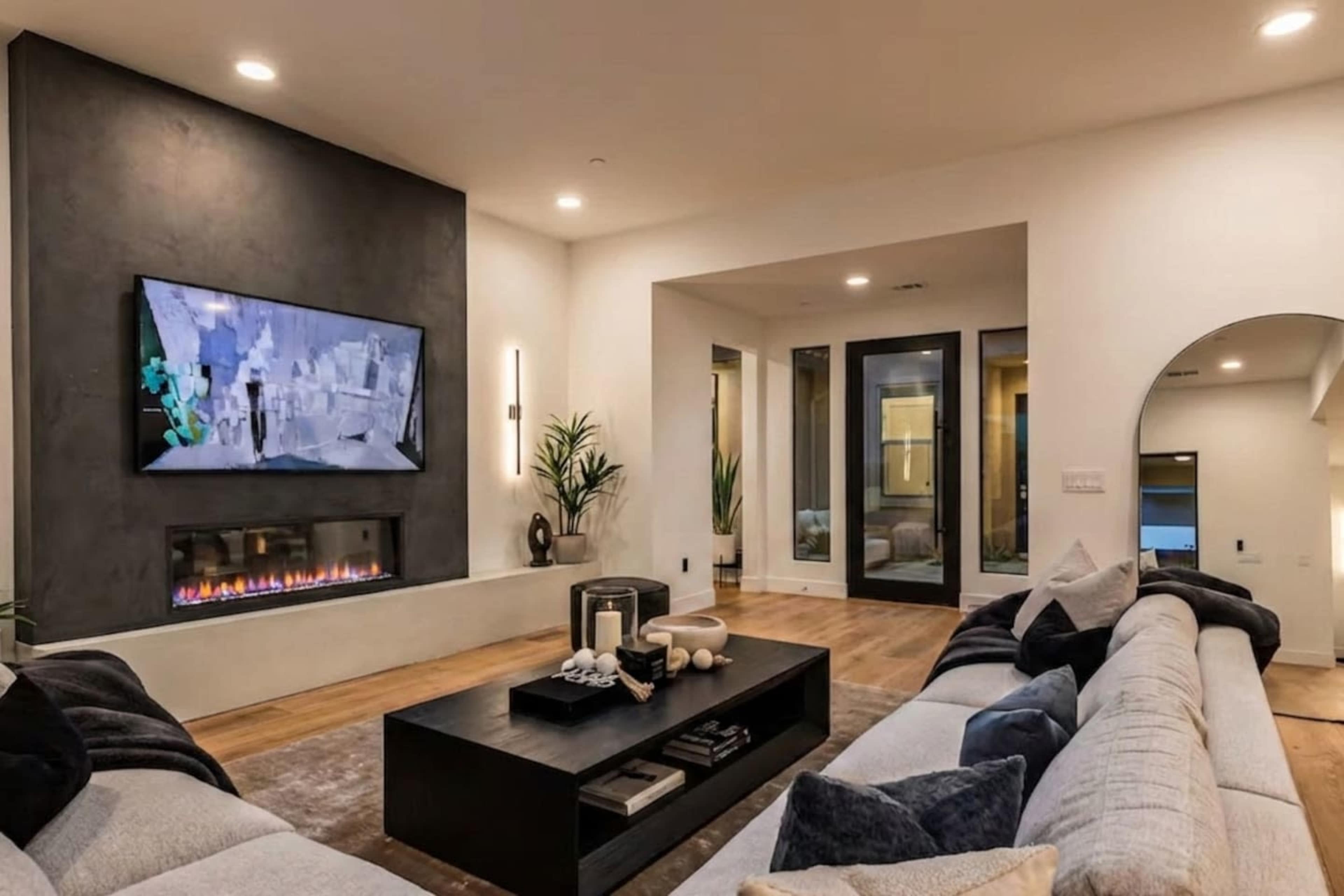 A modern living room features a wall-mounted television above a sleek fireplace, with a central coffee table and a sectional sofa along the perimeter.