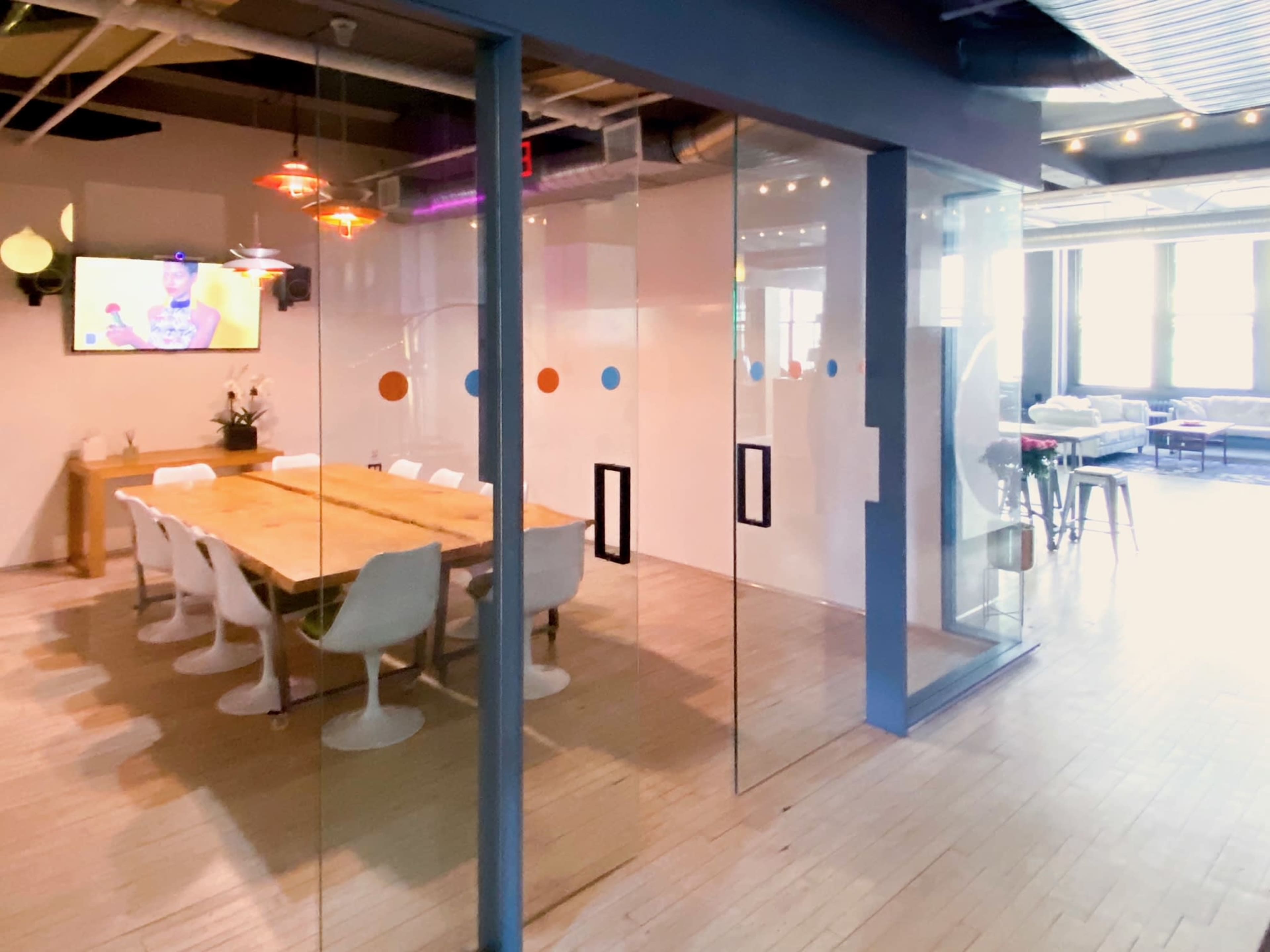 A modern office space with a conference room featuring a long wooden table surrounded by white chairs, separated by glass walls, and visible seating areas in the background.