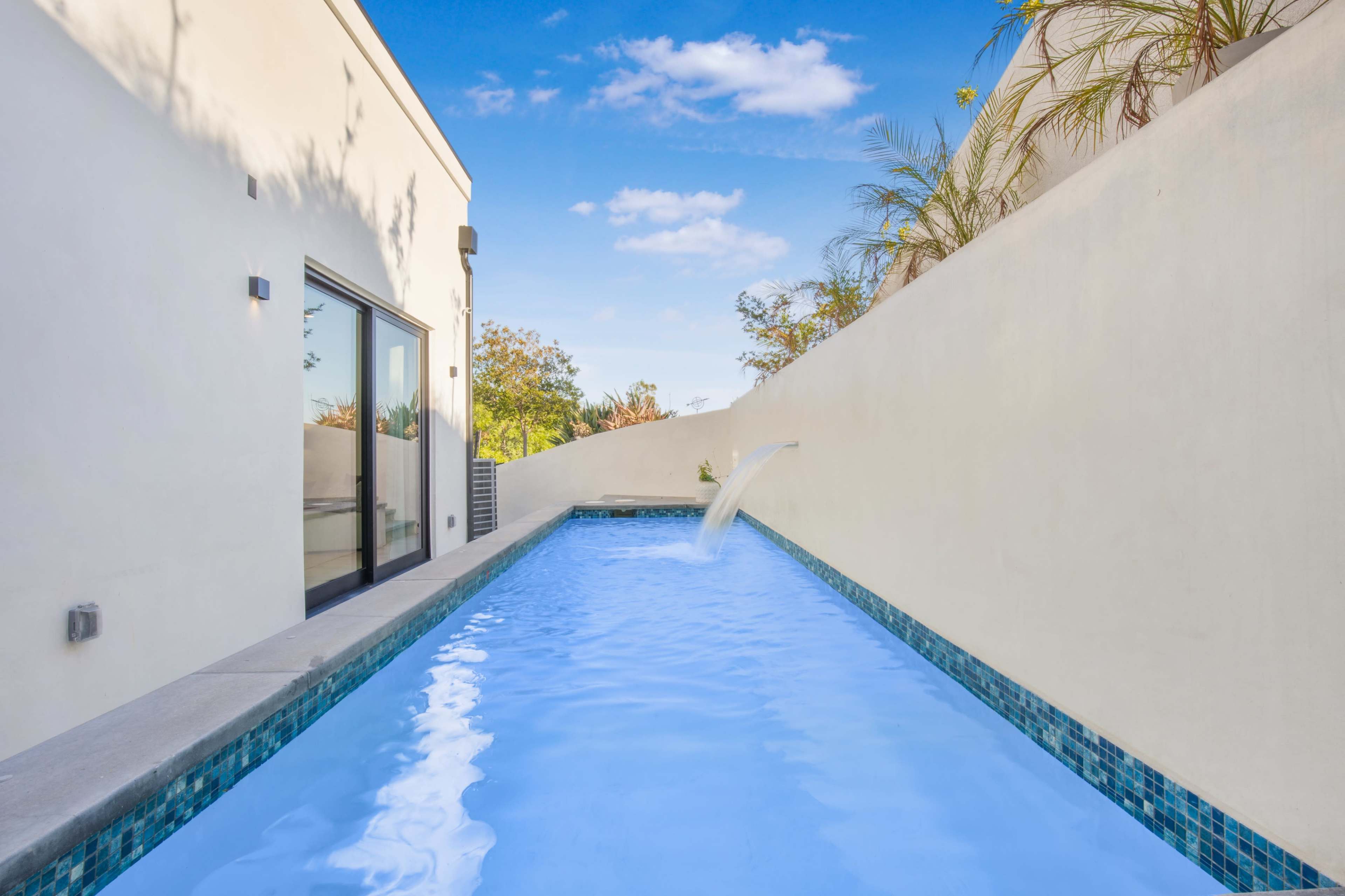 The image shows a modern swimming pool bordered by a clean, white wall and surrounded by greenery under a clear blue sky.