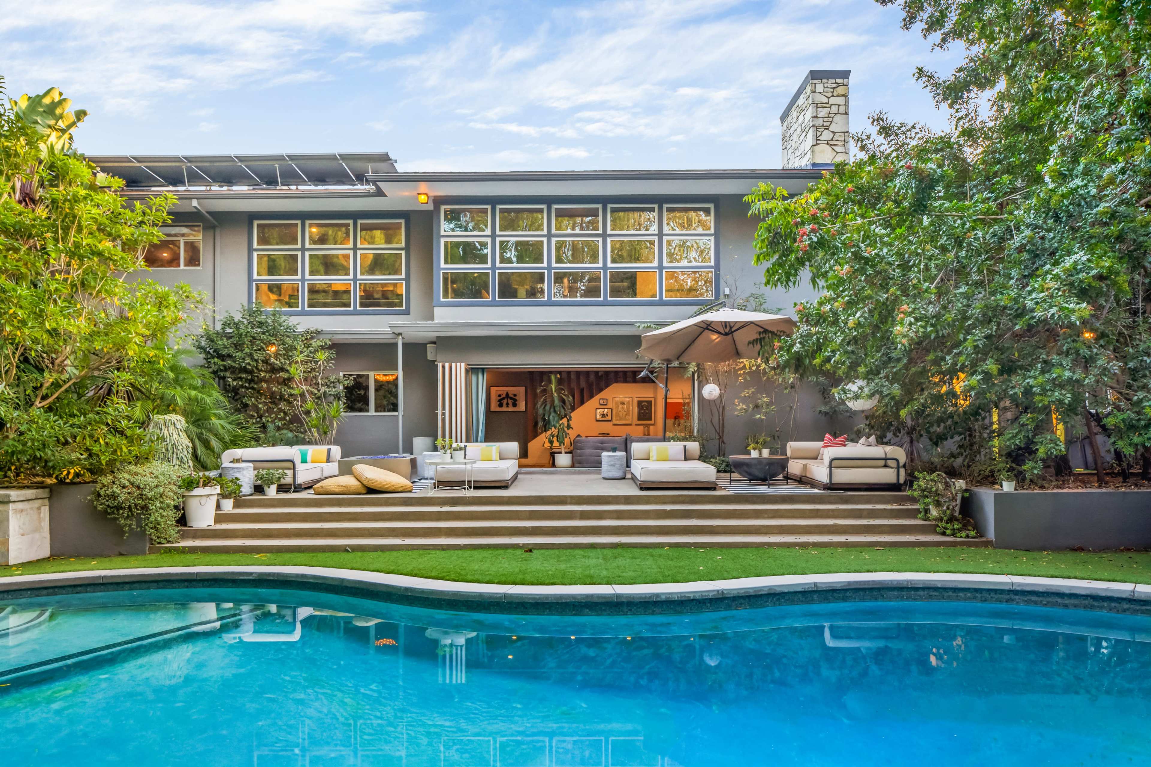 A modern house with large windows overlooks a swimming pool surrounded by lounge seating and greenery.