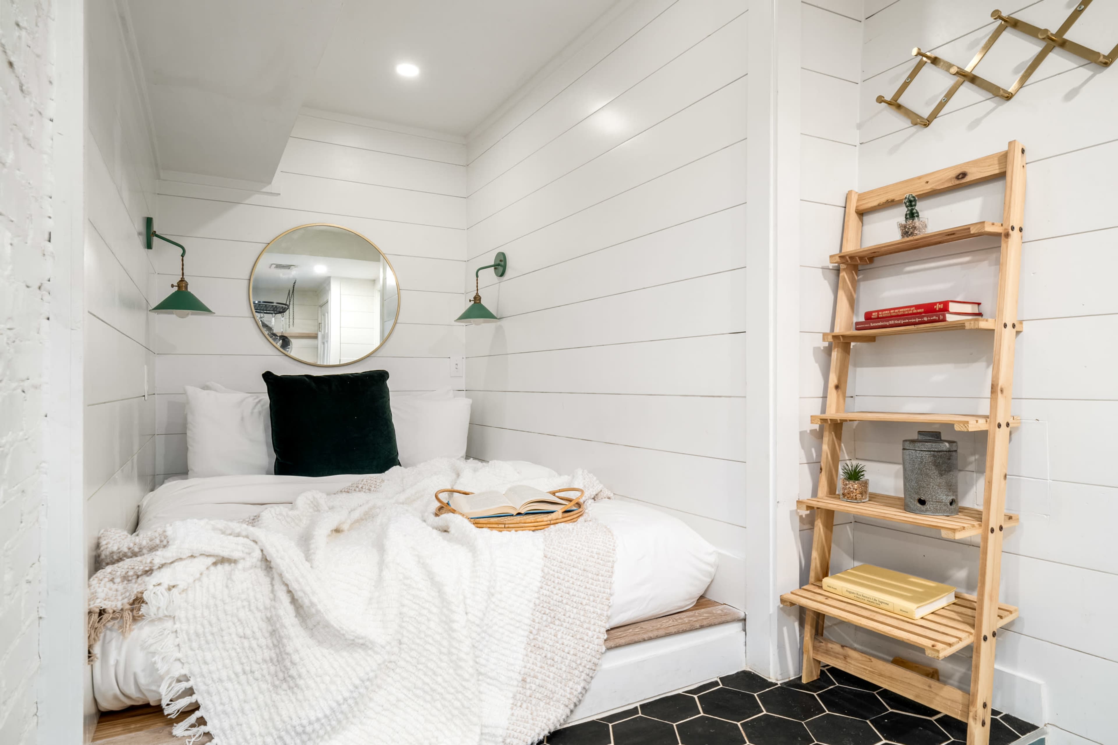 The image shows a small, cozy bedroom with a double bed, white shiplap walls, a round mirror, green wall sconces, and a wooden shelf.