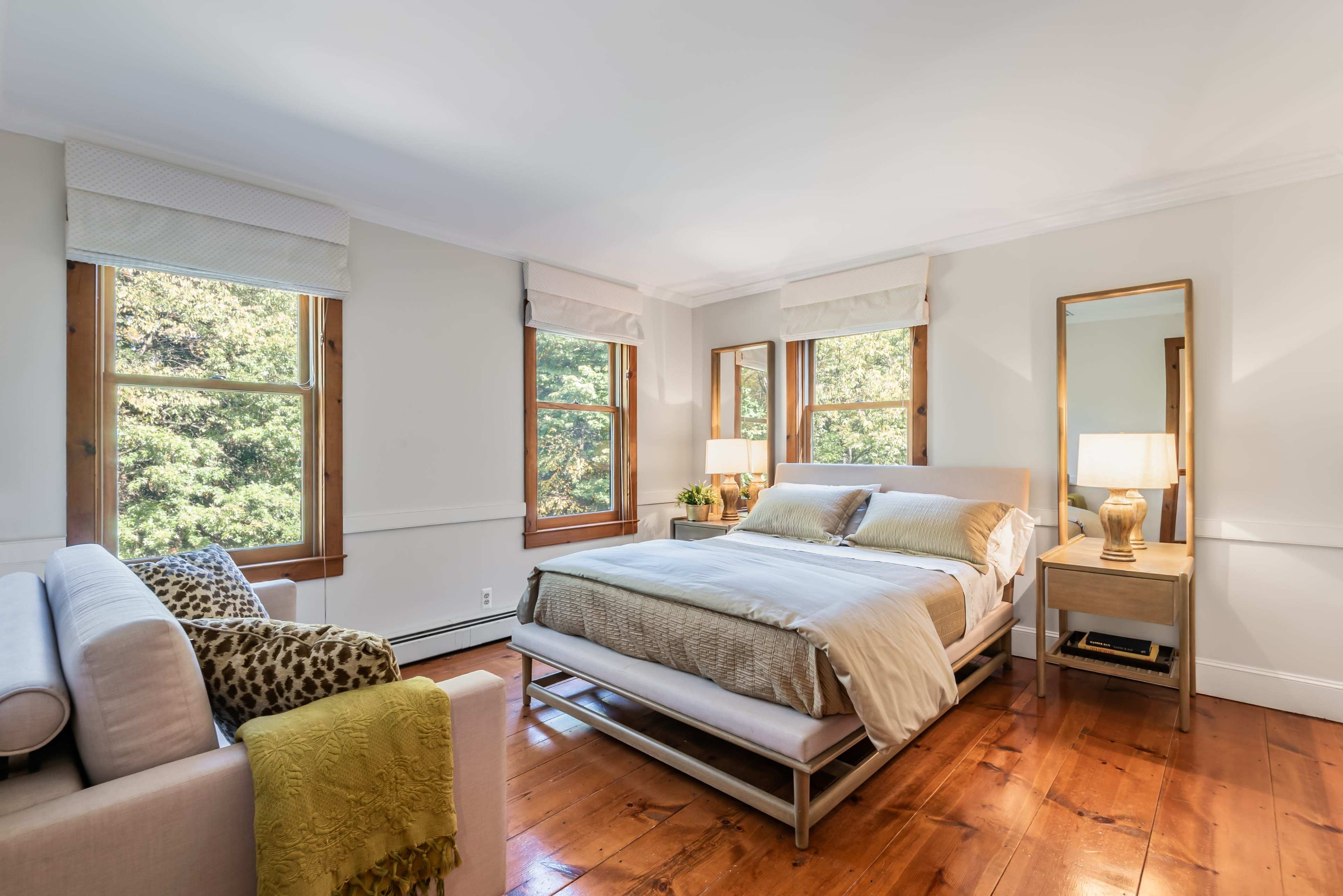 A well-lit bedroom featuring a bed with neutral bedding, two windows allowing natural light, a sofa, a nightstand with a lamp, and wooden floors.