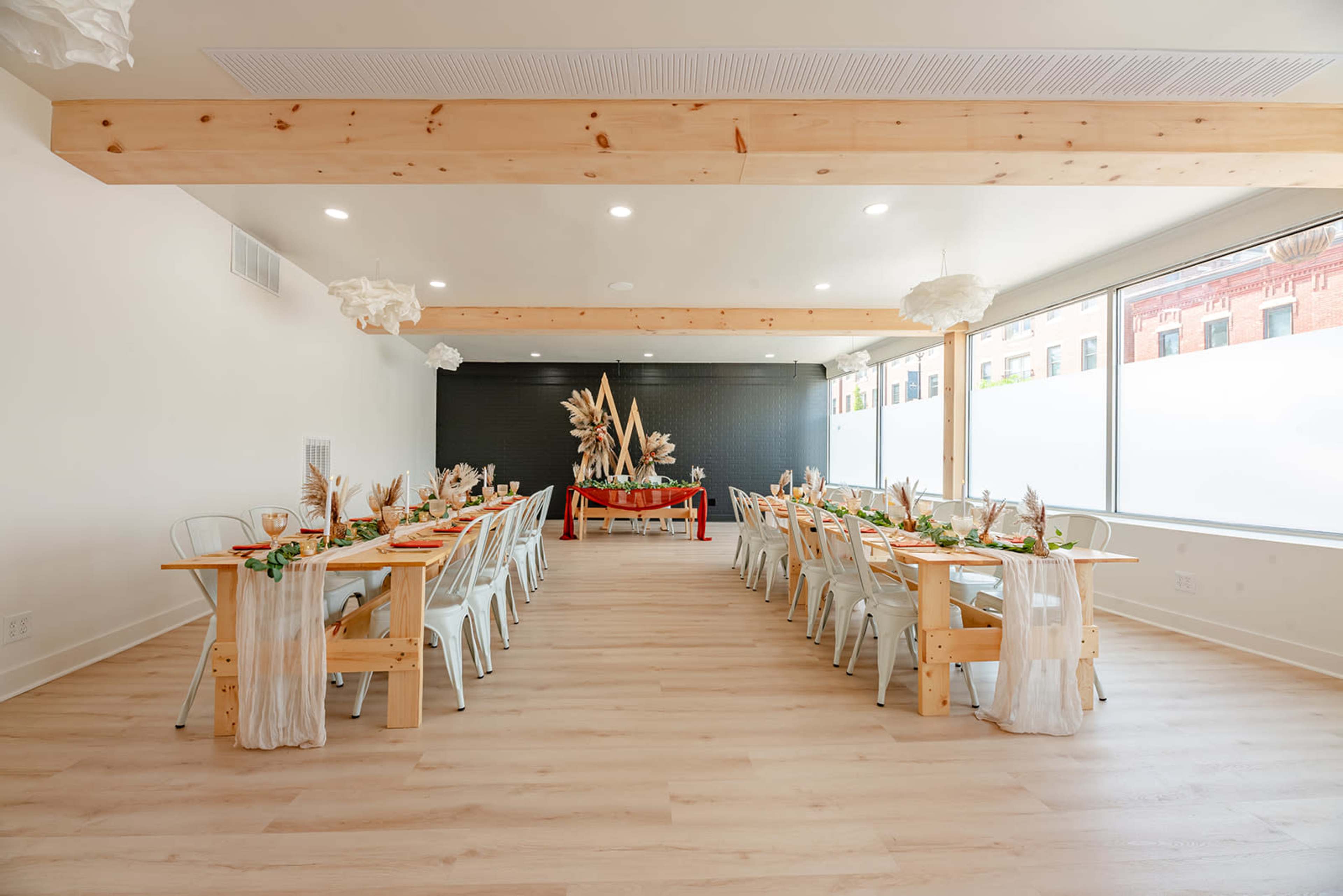 A modern dining room features long wooden tables set for a meal, with a decorative centerpiece at the far end and large windows providing natural light.
