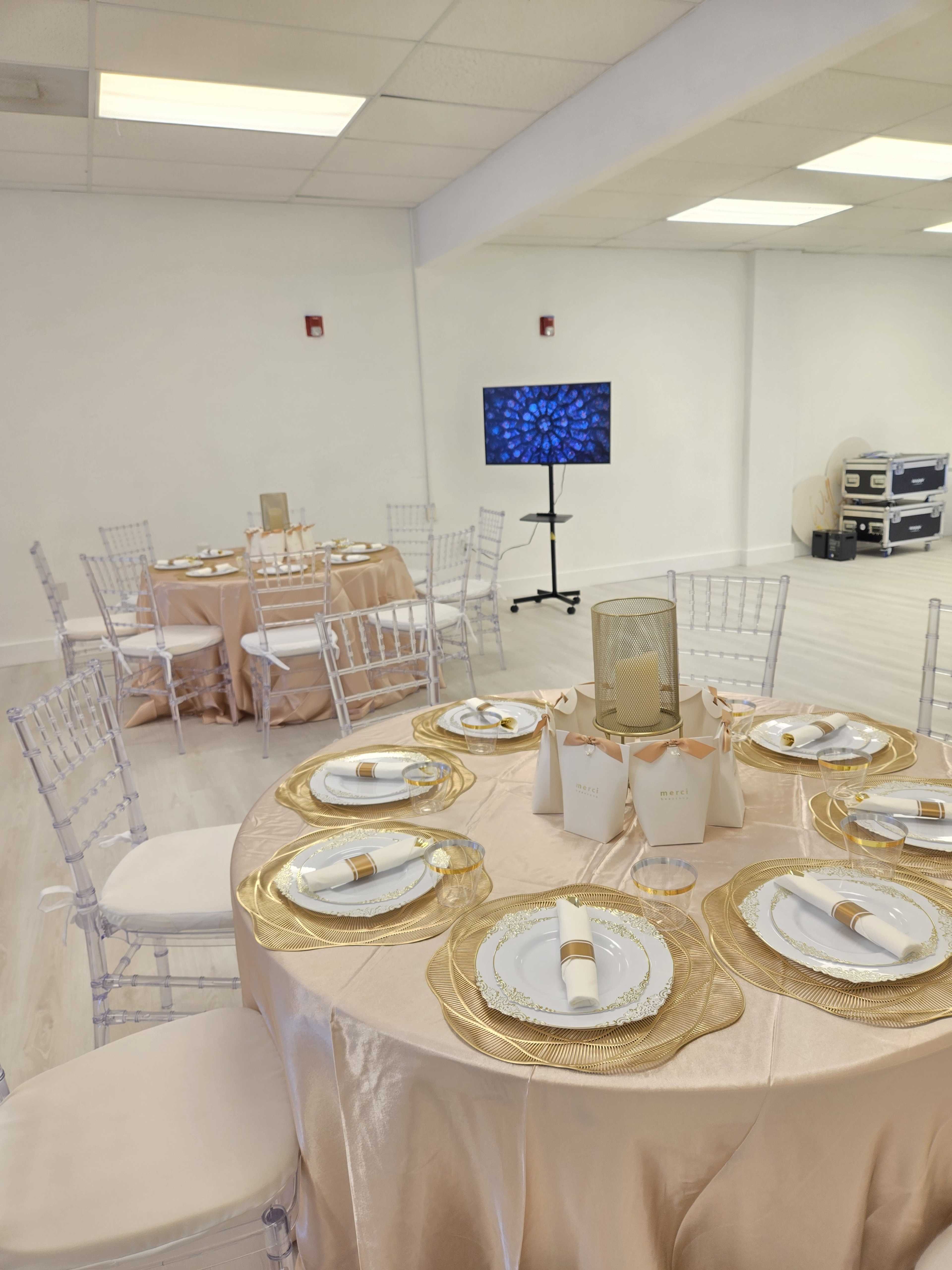 A well-decorated event space with elegantly set tables featuring gold and white tableware, a central decorative piece, and a television displaying a pattern in the background.